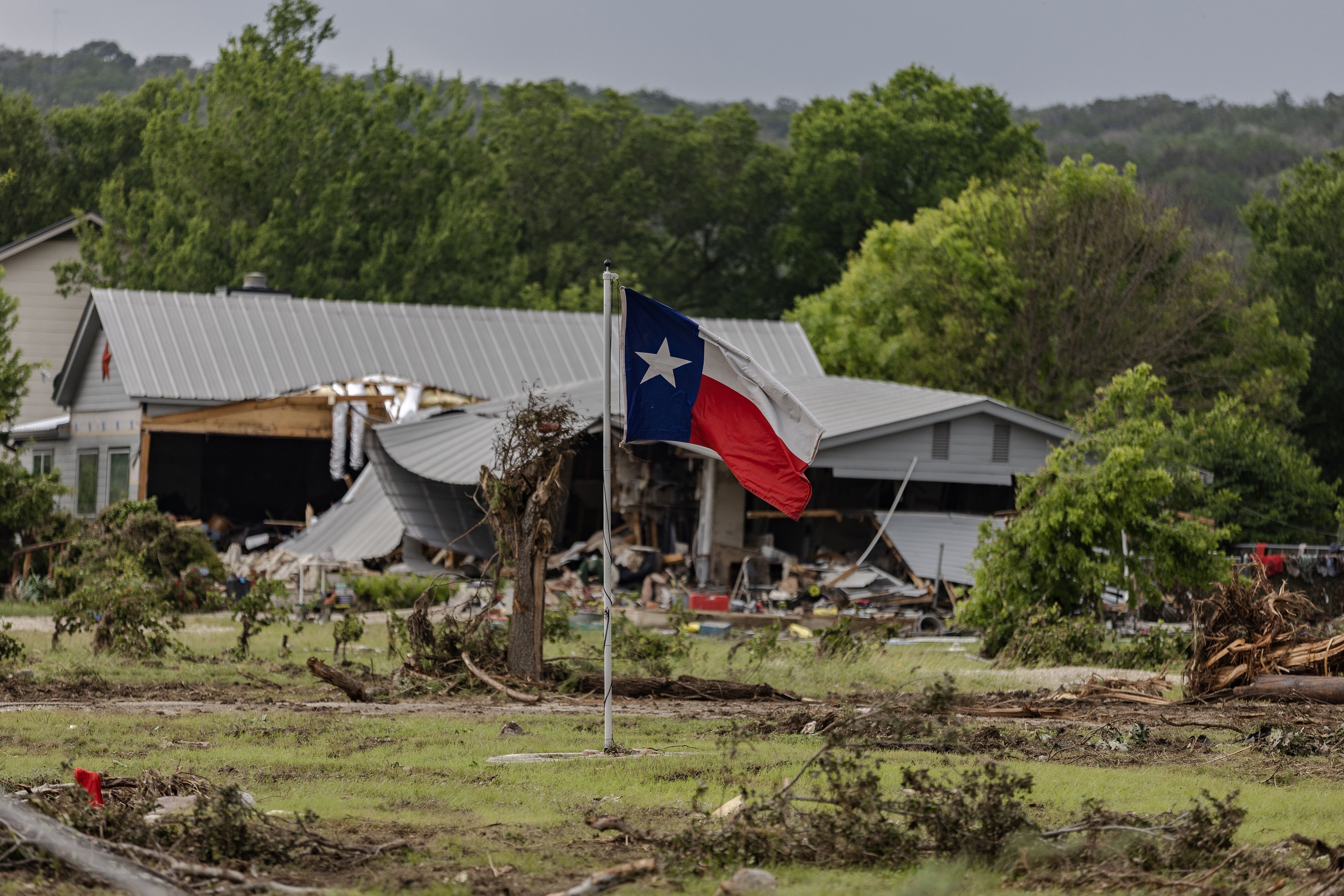 Texas flag on a pole in front of a wrecked gray metal-roofed building with debris and broken trees scattered on a grassy field under a gray sky.
