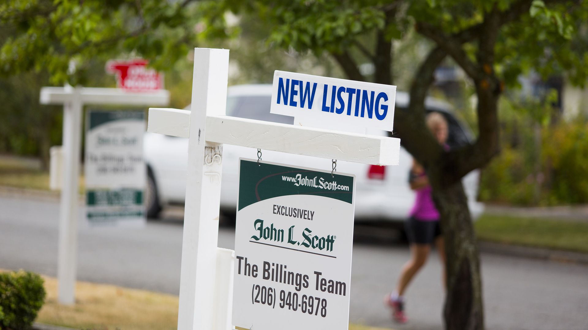 A real estate sign posted on the edge of a street with "New Listing" attached to the top of the sign post. Another for sale sign is visible in background.