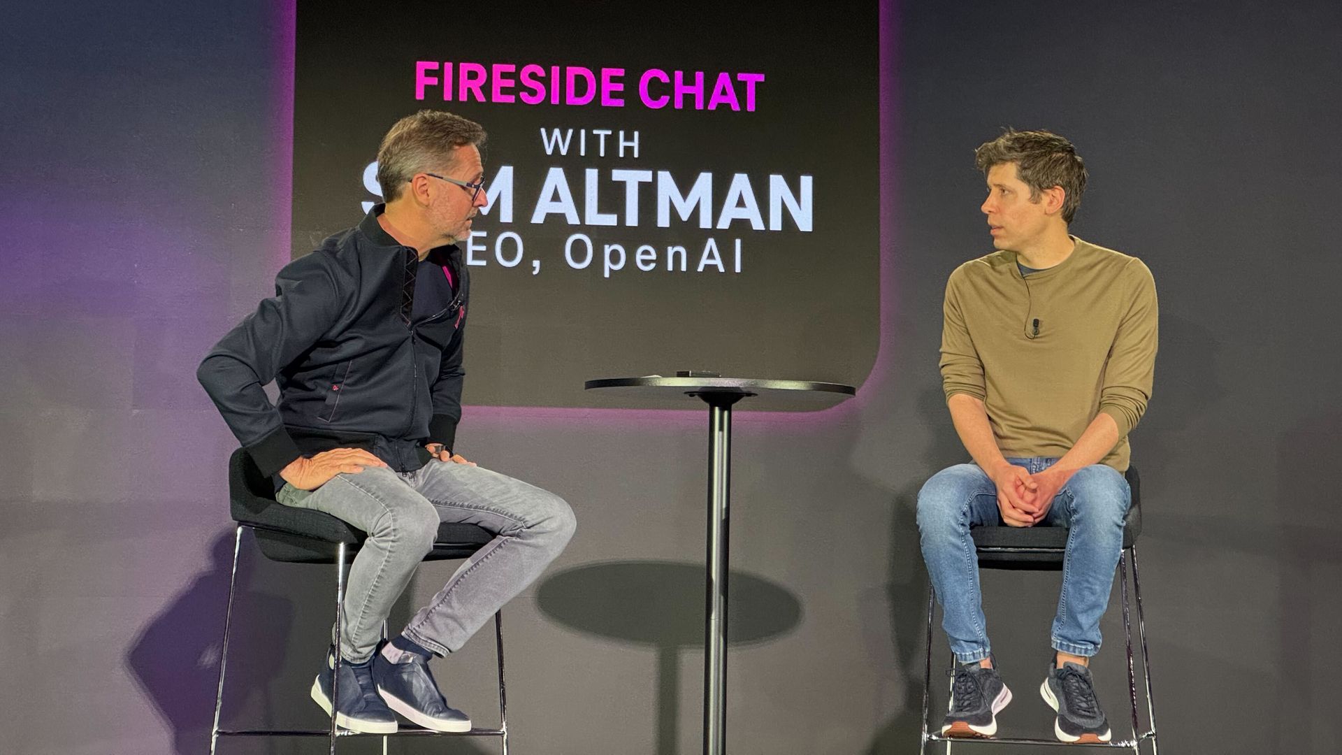 Photo of Sam Altman in sweatshirt and blue jeans on a stool on stage being interviewed by T-Mobile CEO 