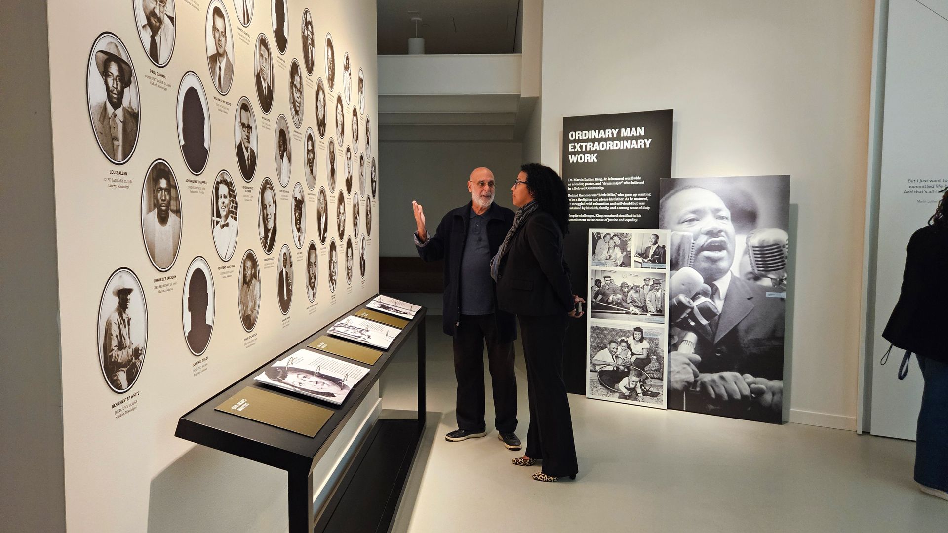 Two people discuss a museum exhibit featuring black-and-white portraits of historical figures and a large image of Martin Luther King Jr. with the title "Ordinary Man Extraordinary Work."