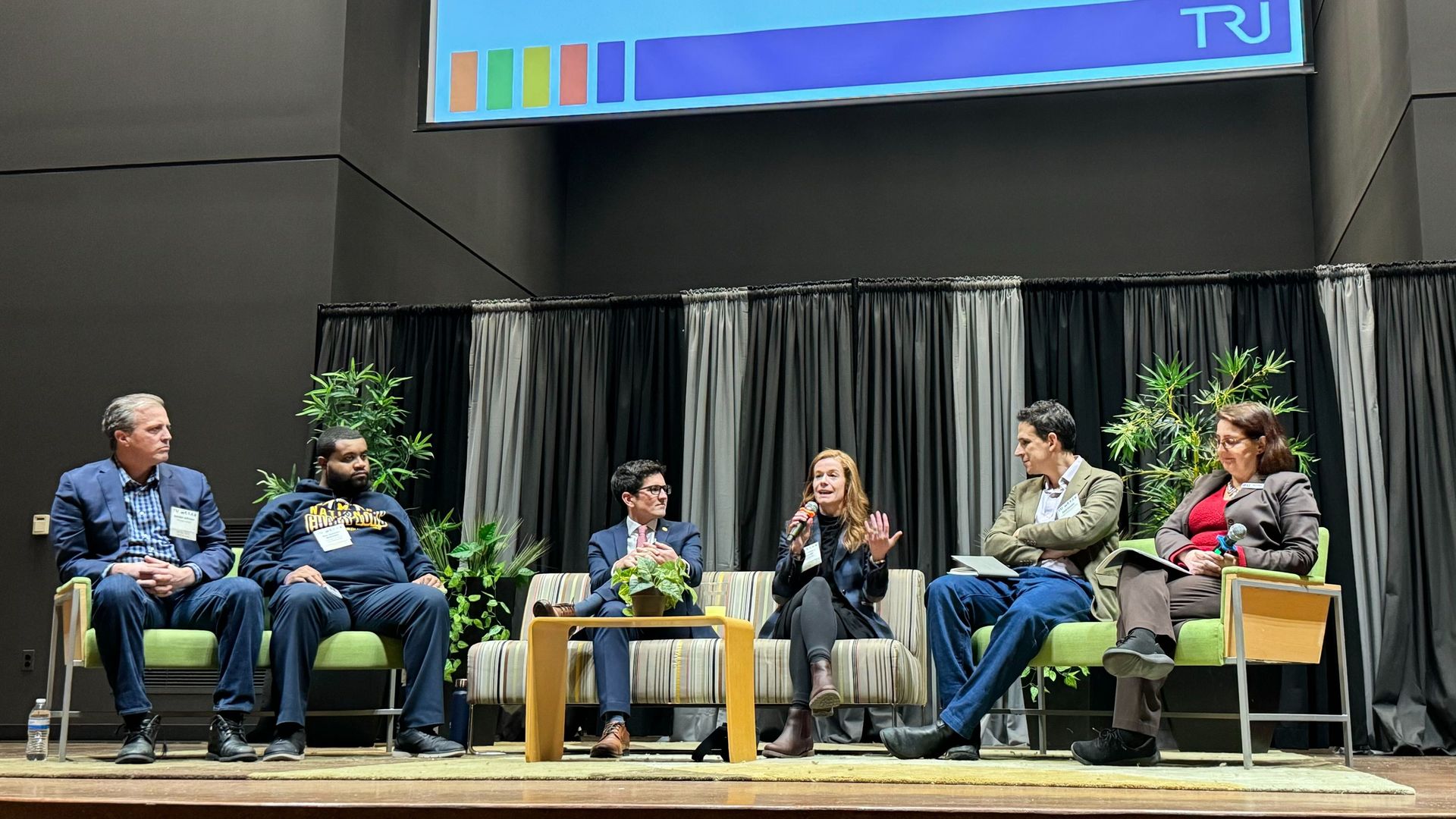 Policymakers on stage listen as woman discusses transit while gesturing.