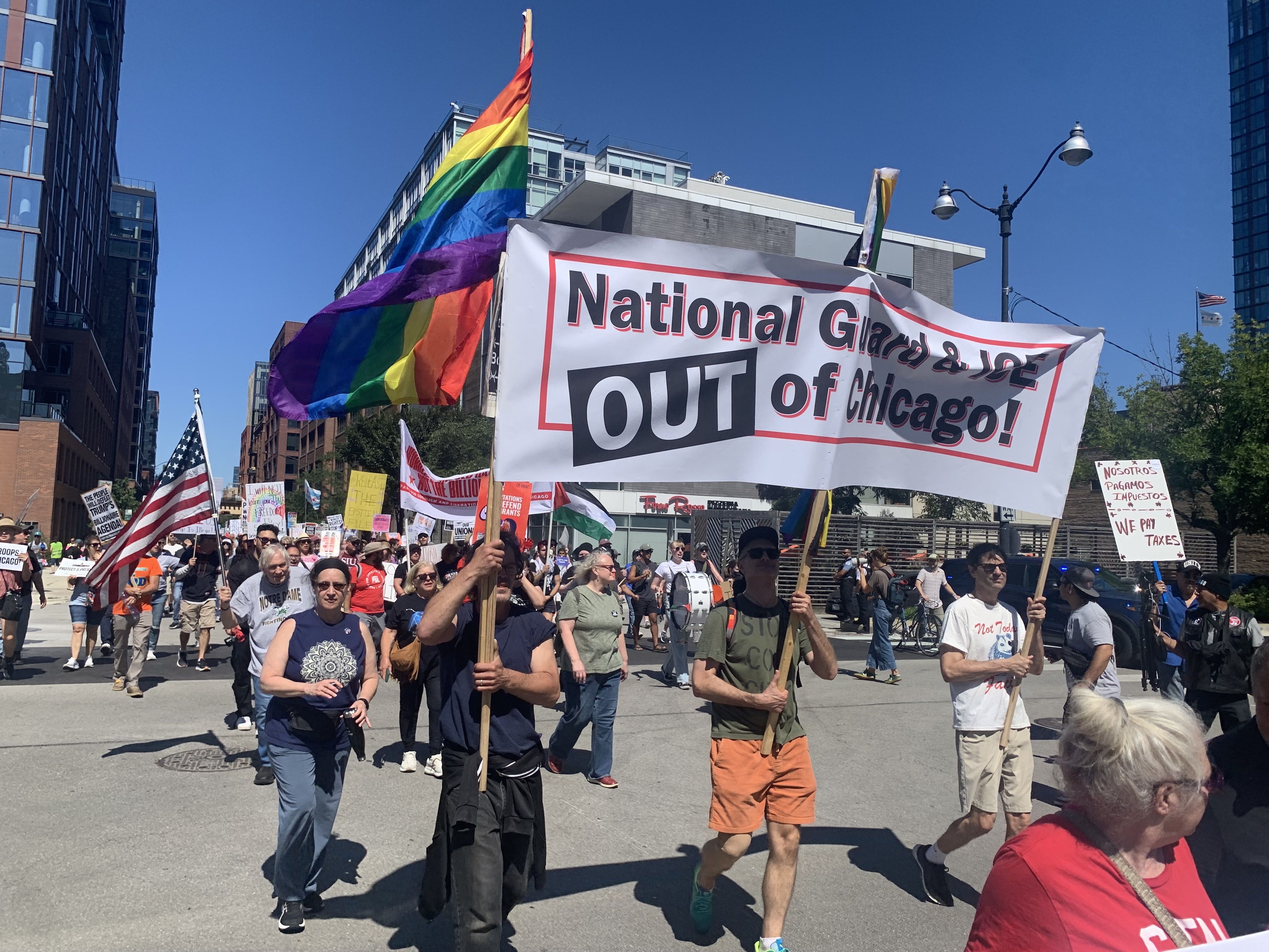Protesters march on a sunny city street holding signs, including a large banner saying "National Guard & JOE OUT of Chicago!" and a rainbow pride flag waving.