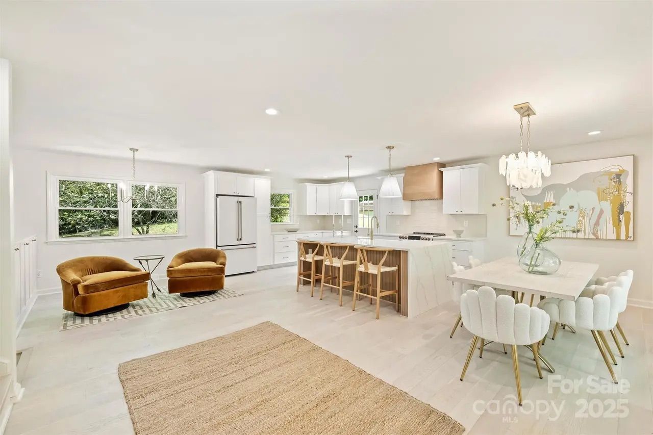Bright open kitchen and dining area with white cabinetry, marble island, wooden stools, beige rug, two brown velvet chairs by window, white dining chairs with gold legs, modern chandelier.