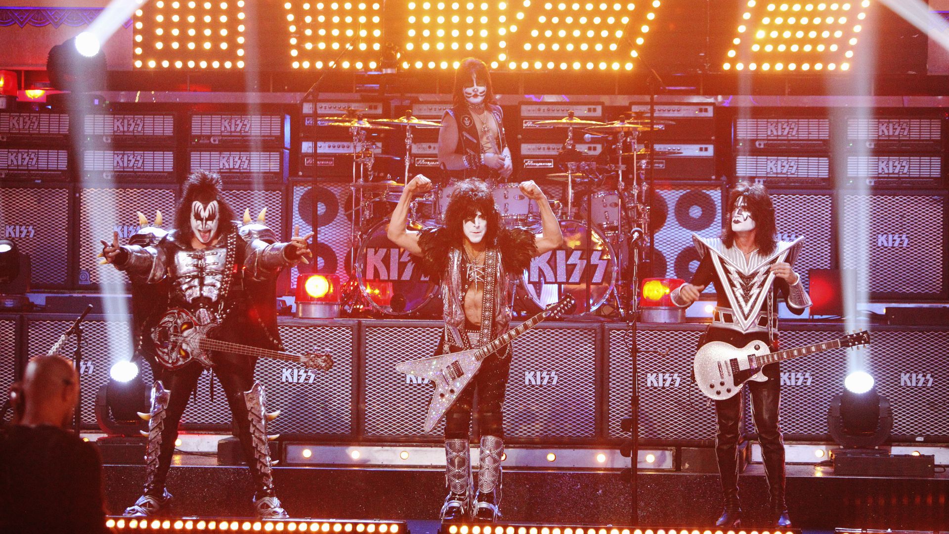 Three rockers in black and white face paint hold guitars while performing on a stage