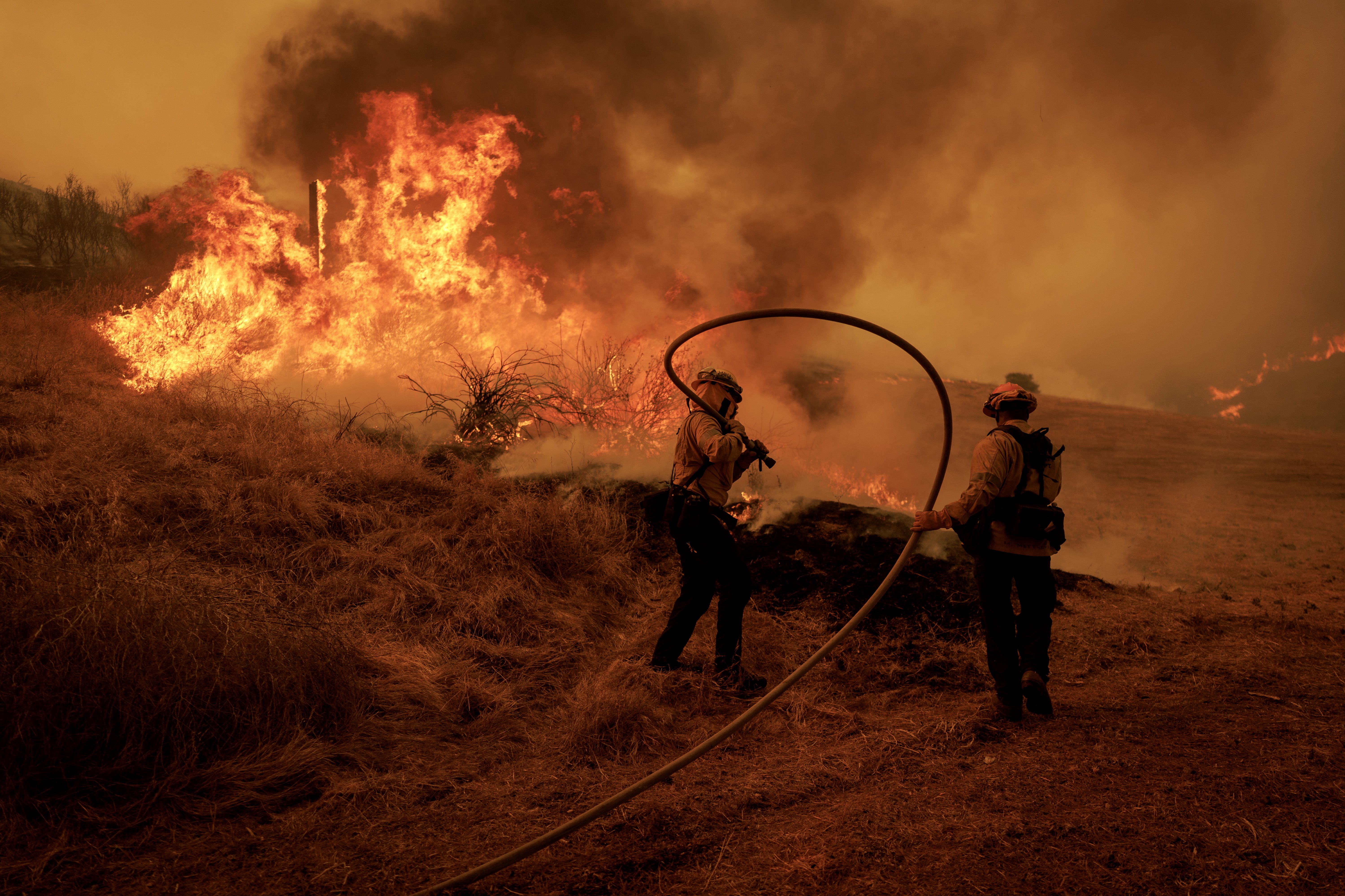 Two firefighters with helmets hold a fire hose as they battle a large wildfire in dry, grassy terrain with intense orange flames and thick smoke.