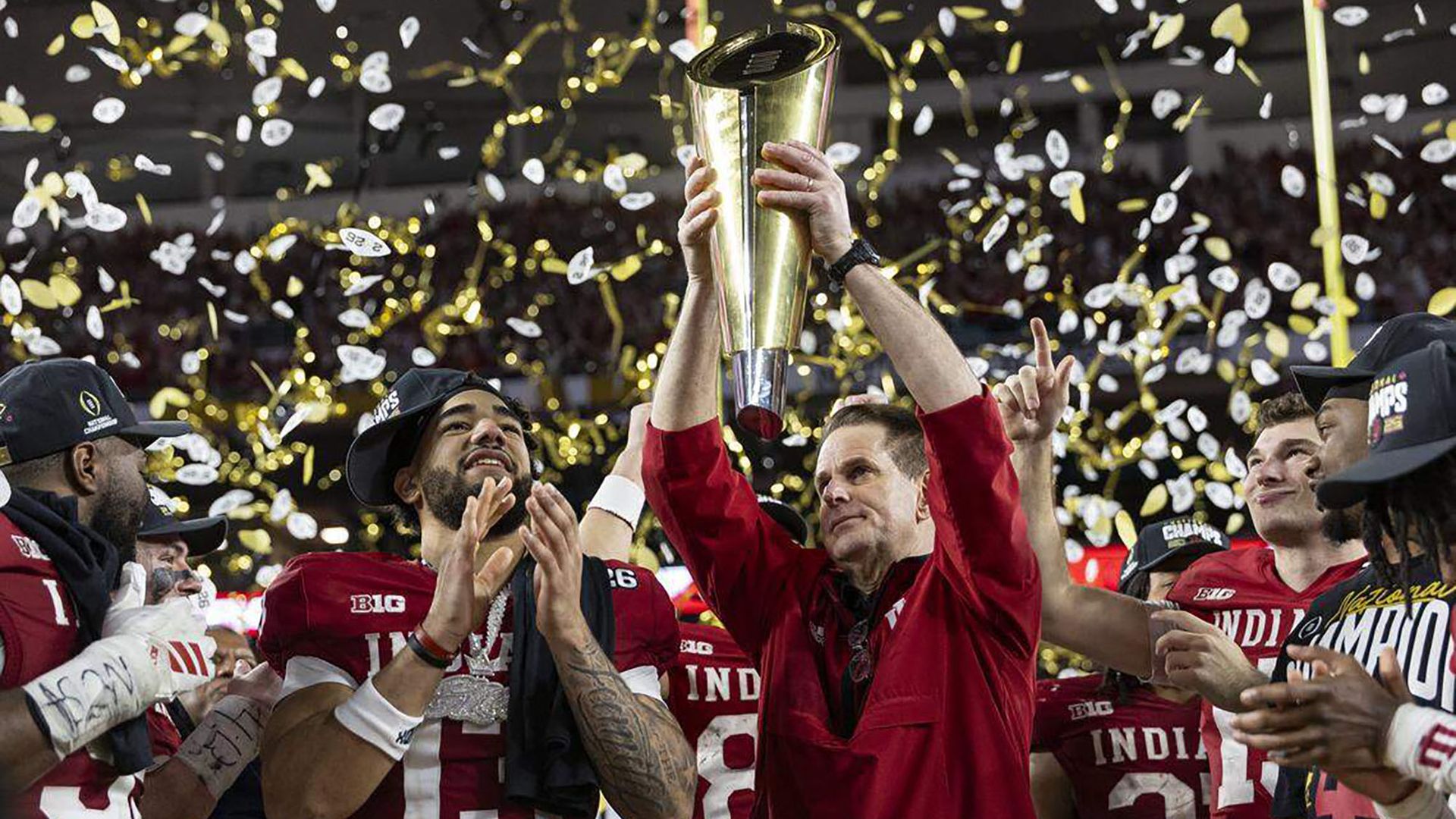 Indiana football players celebrate, with the coach raising a tall gold trophy high as gold and white confetti falls; red jerseys spell INDIANA while teammates cheer nearby.