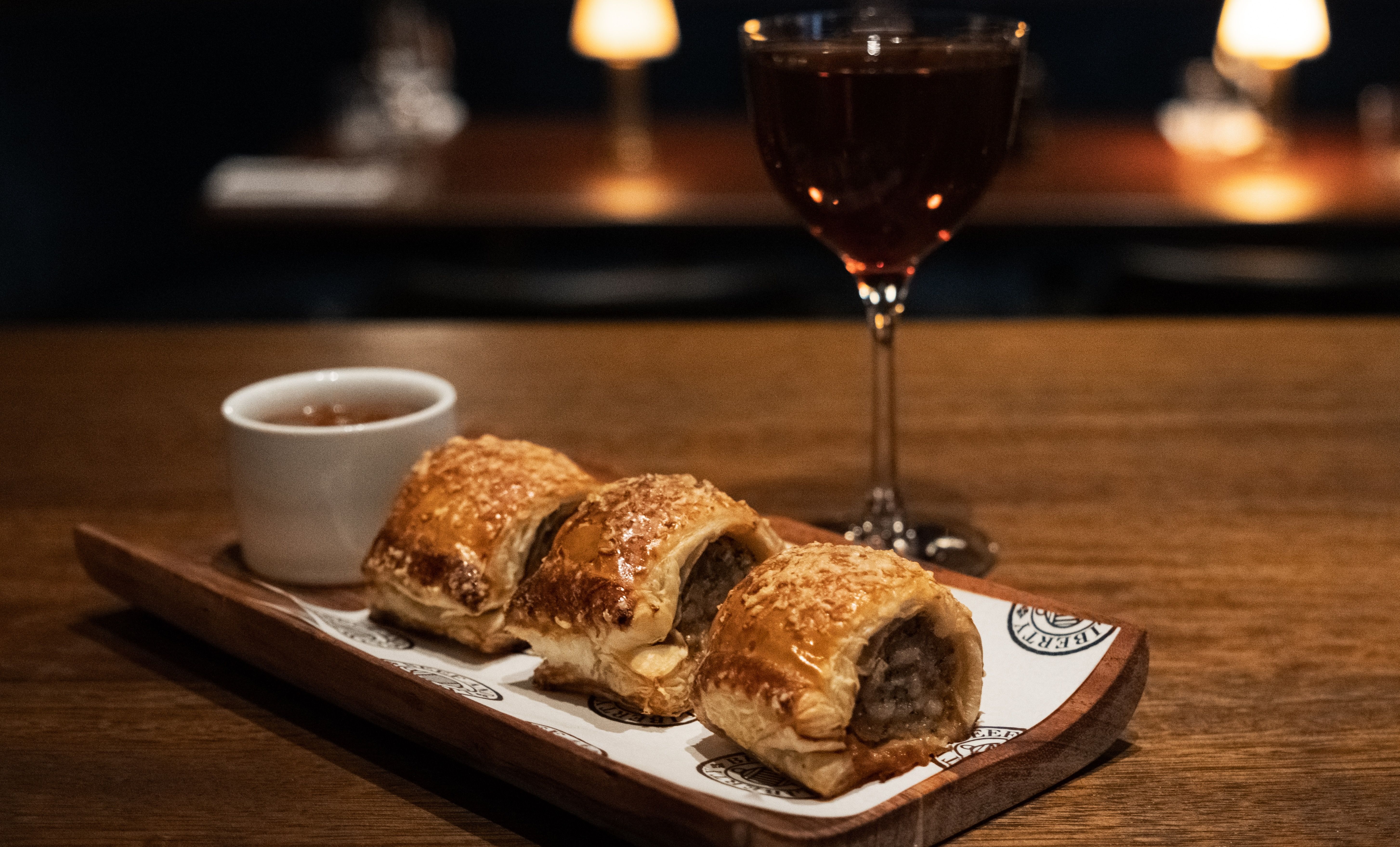 Three golden-brown sausage rolls on a wooden tray with a small white cup of dipping sauce and a filled wine glass in a dimly lit setting.