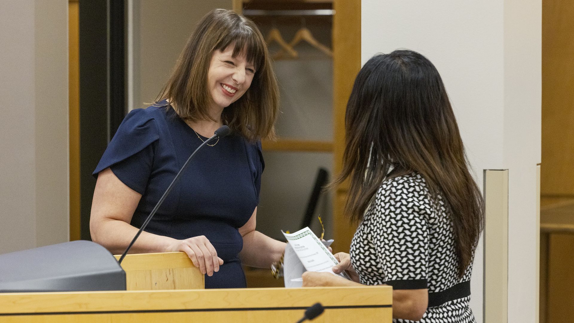 Two women at a wooden podium: one in a navy dress smiles as she hands a certificate to the other in a black-and-white patterned dress; a microphone sits on the lectern.