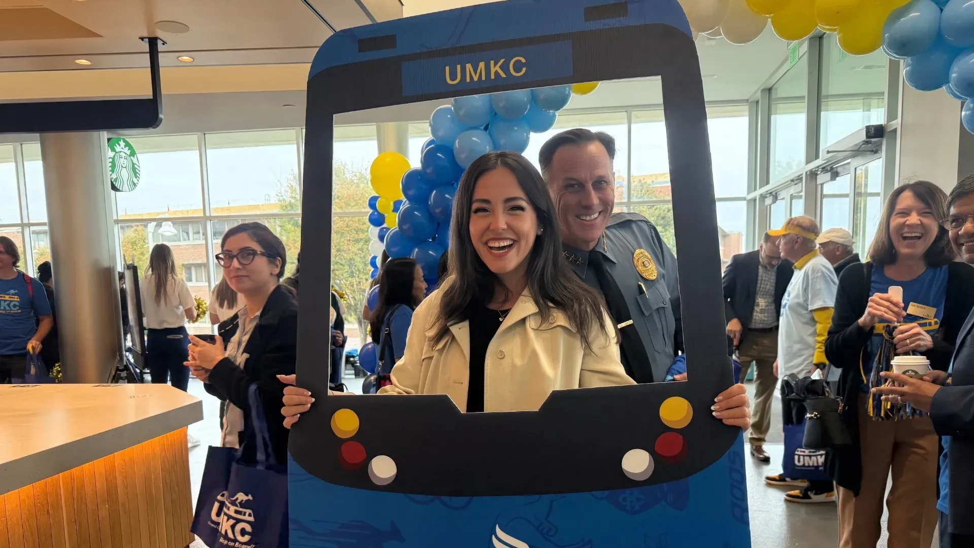 Two people smiling and posing behind a blue and black UMKC-themed cutout, inside a room with windows and blue and yellow balloon decorations, with several people in the background.