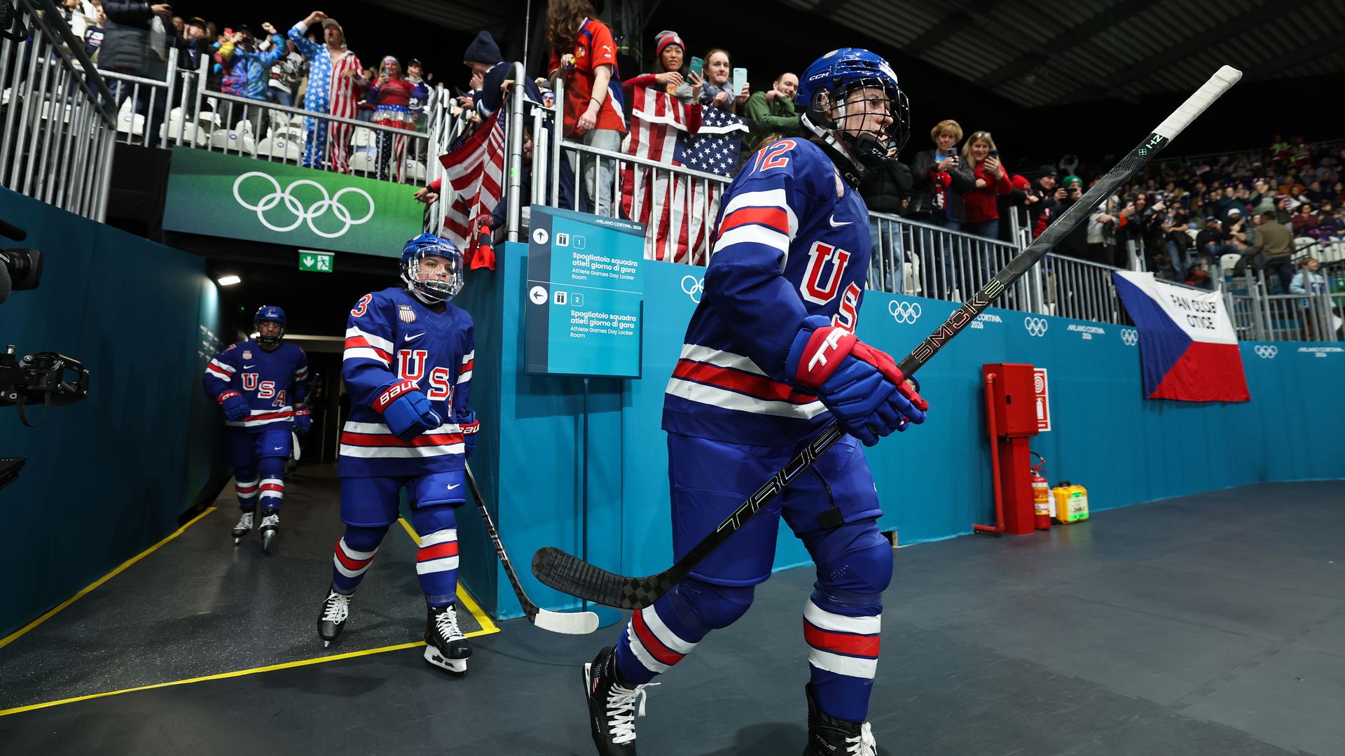 USA women's ice hockey players in blue uniforms with red and white stripes enter the rink, holding sticks, while fans dressed in USA apparel cheer from above at the Olympics venue.