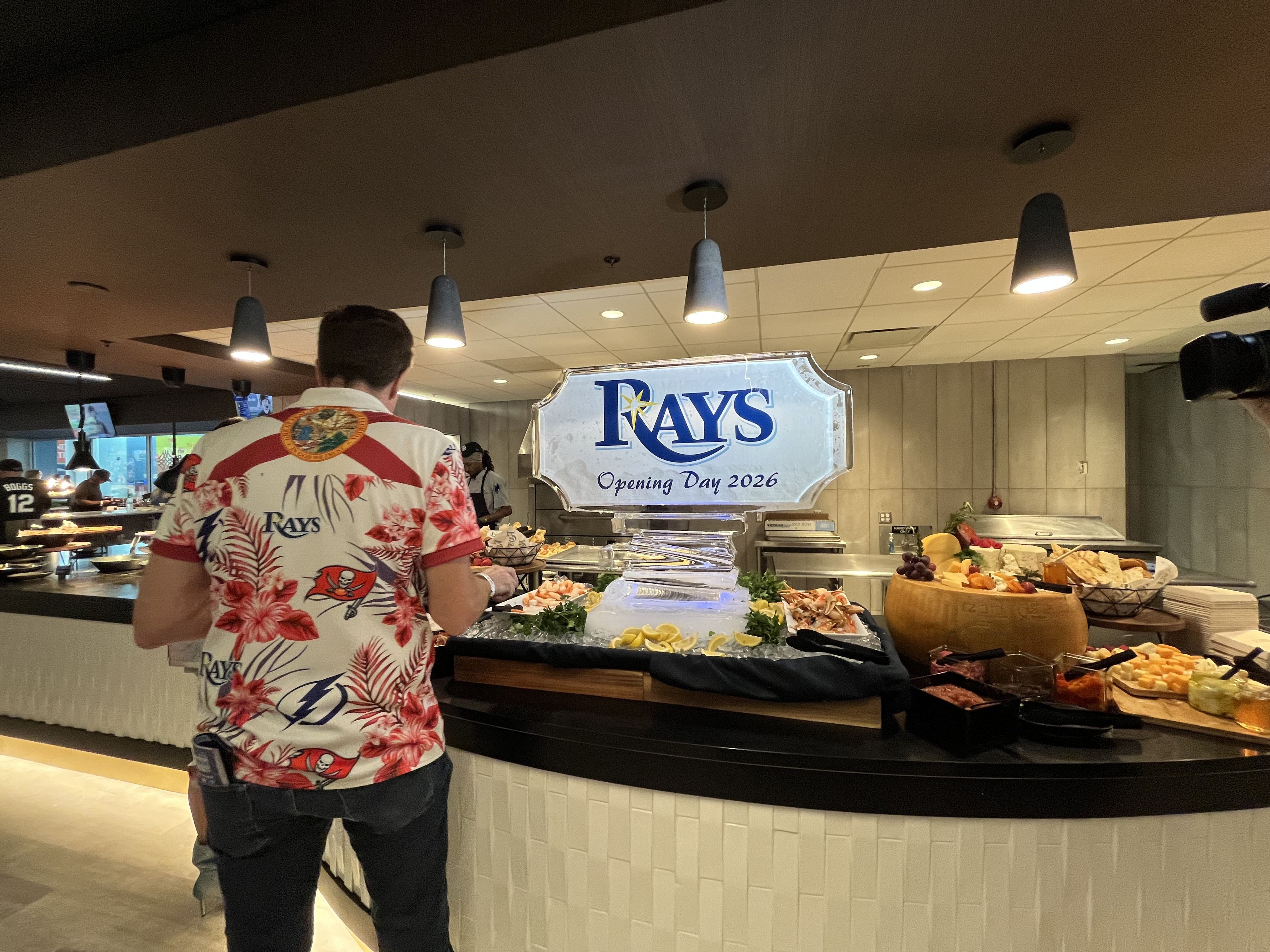A man wearing a tropical Rays shirt stands at a curved buffet with cheese, fruit, and bread, beside a large sign that reads "Rays Opening Day 2026" as a camera rolls in the background.