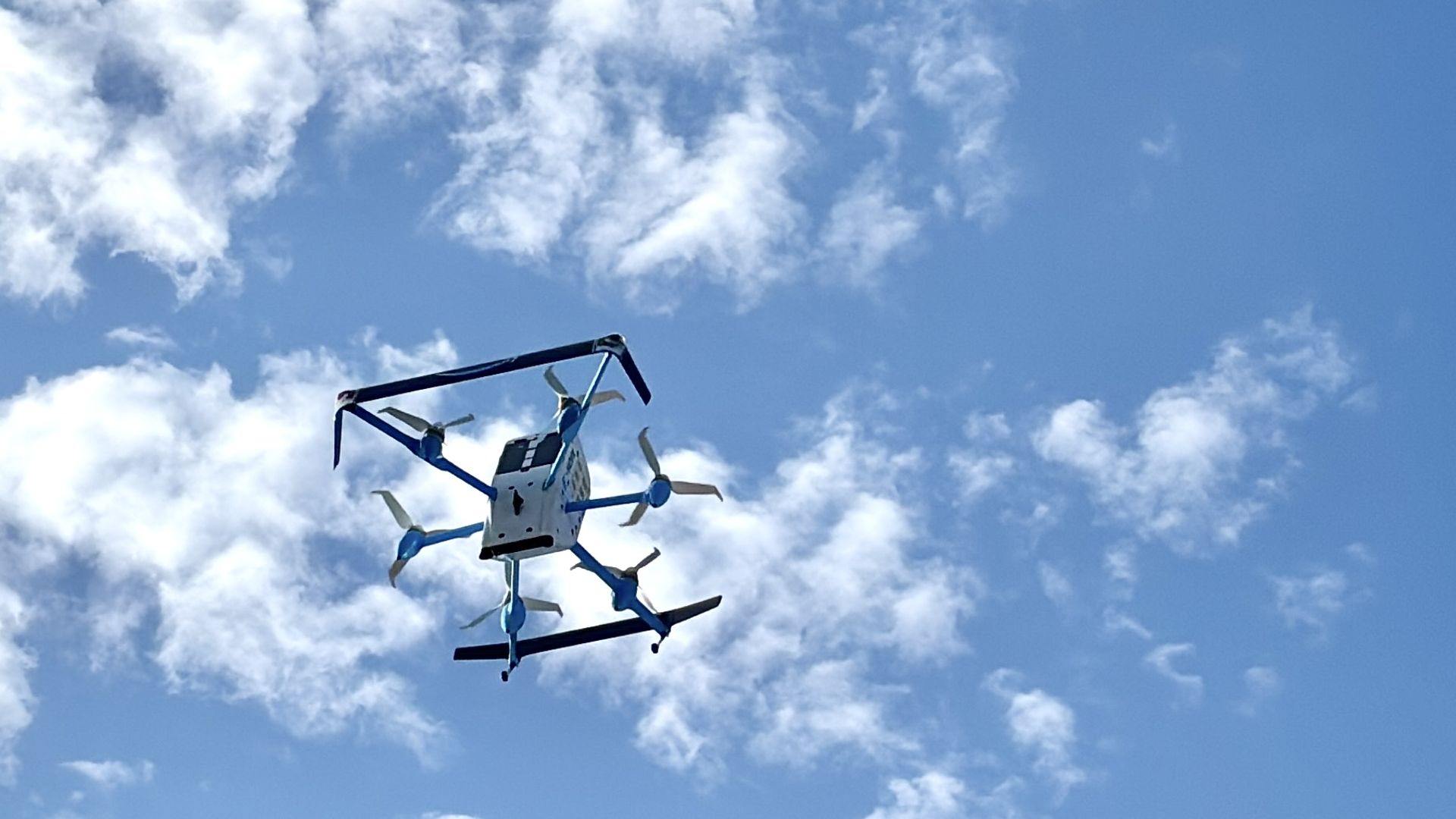 Large Amazon drone six rotors, a white body and blue arms, hovering in a bright blue sky with scattered white clouds.