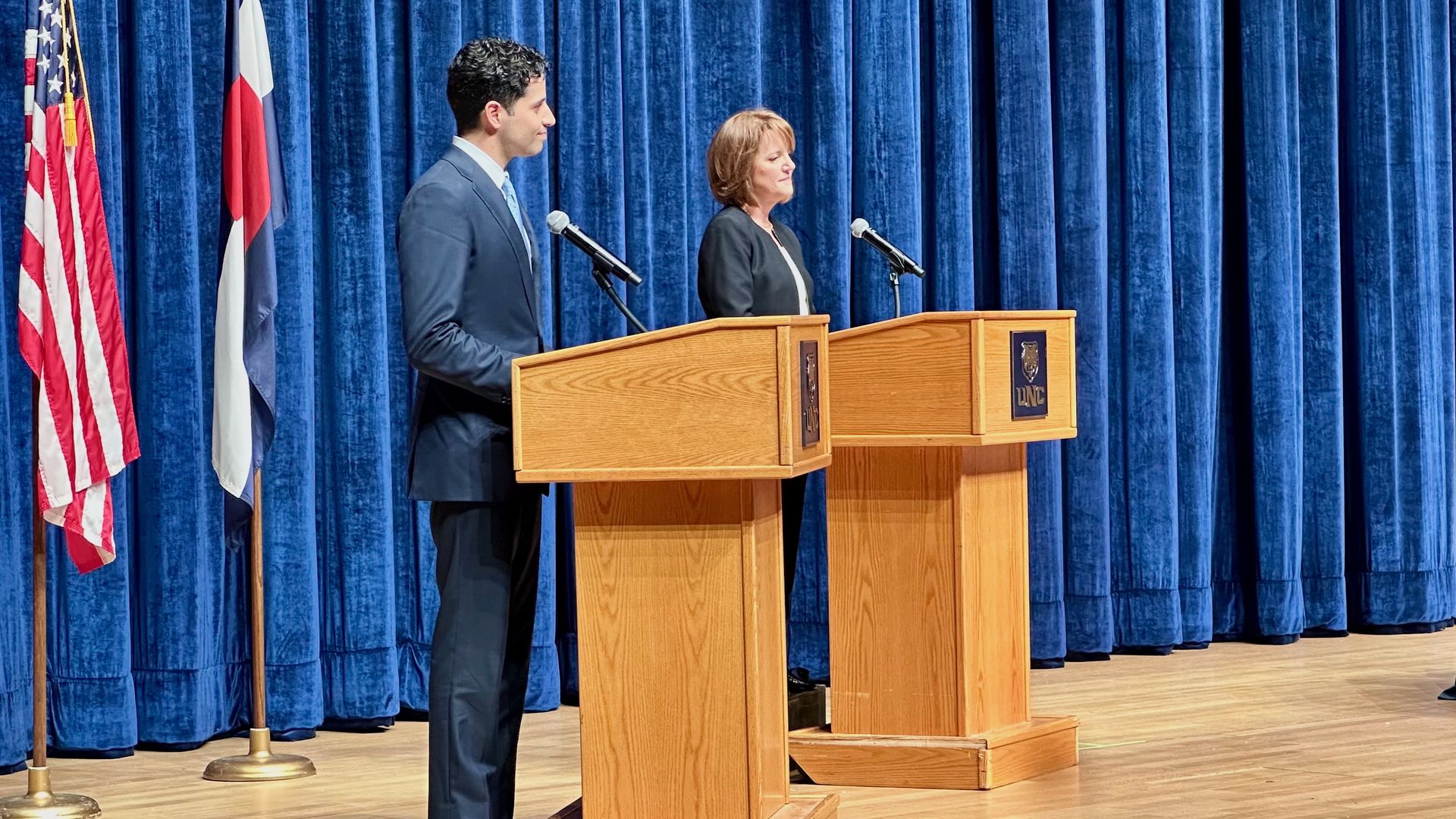 Two speakers, a man and a woman, stand at wooden podiums on a stage with deep blue curtains. Flags stand to the left; a man sits at a table with a microphone and water bottle on the right.