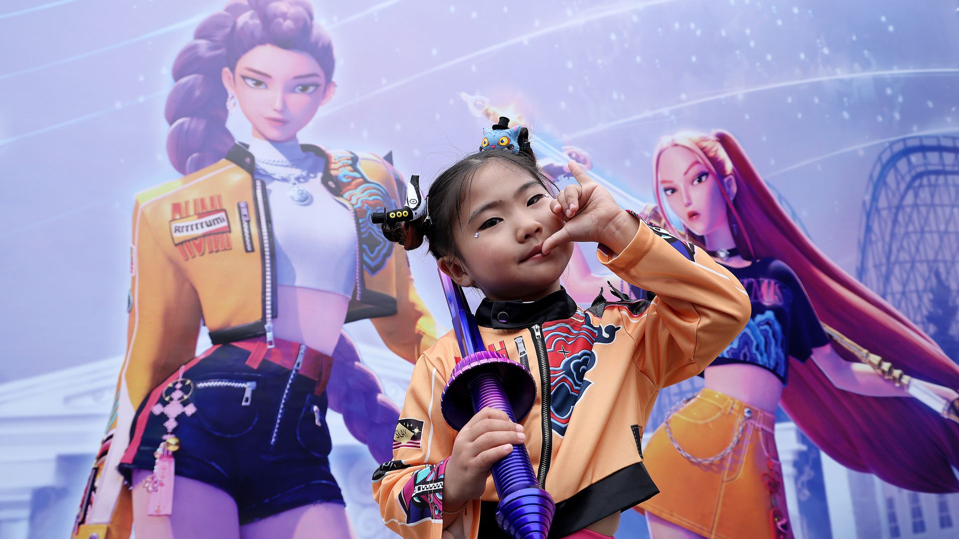 Small girl in orange jacket and blue pants holds a purple toy sword, posing in front of a large poster featuring two animated  characters in colorful outfits.