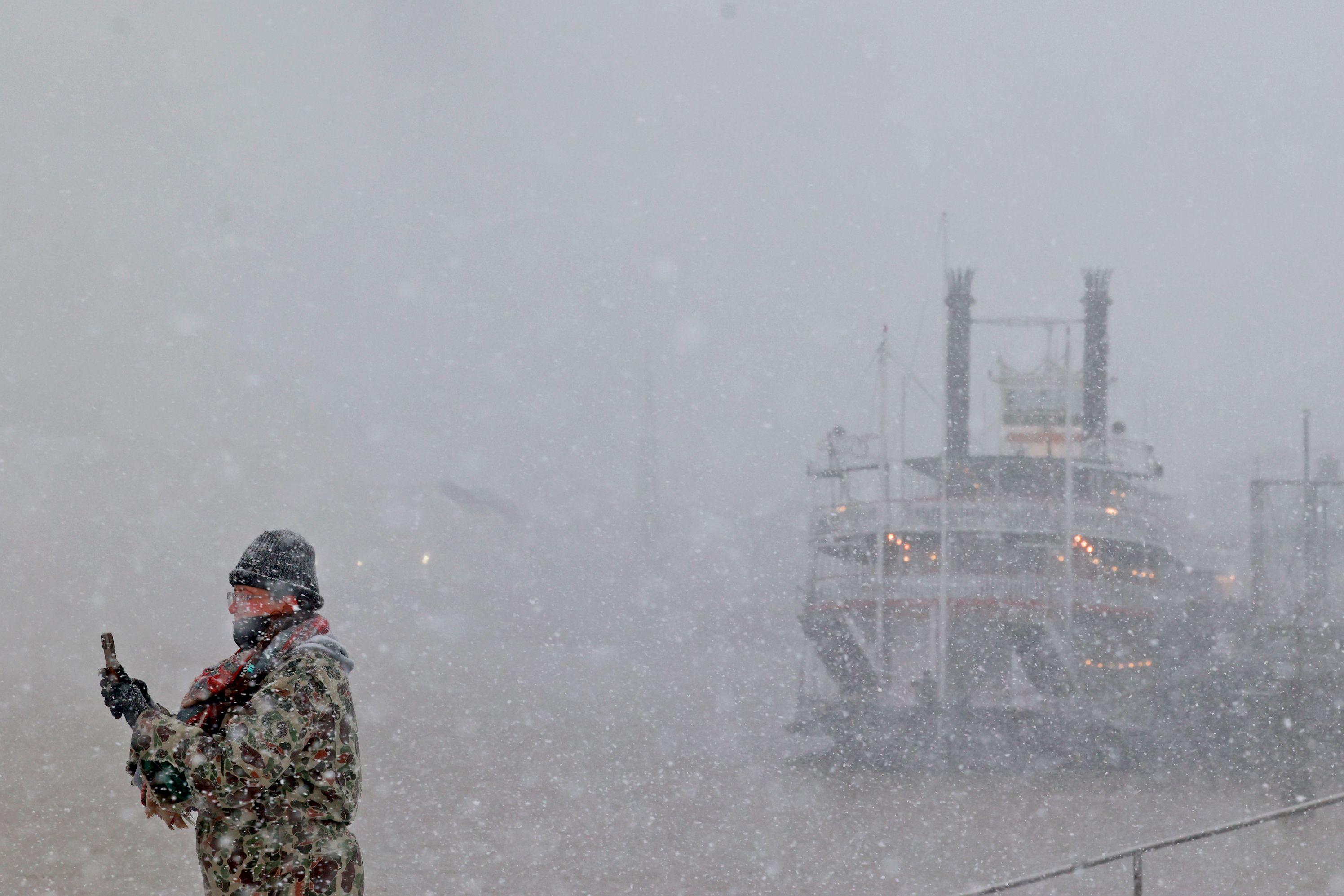 A person takes a cell phone photo as snow falls around them near the Mississippi riverfront.