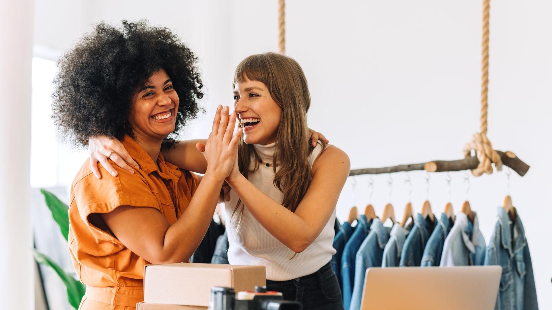 Two smiling women high-fiving in a bright clothing store, one wearing an orange outfit, the other a white sleeveless top, with denim jackets hanging behind them.
