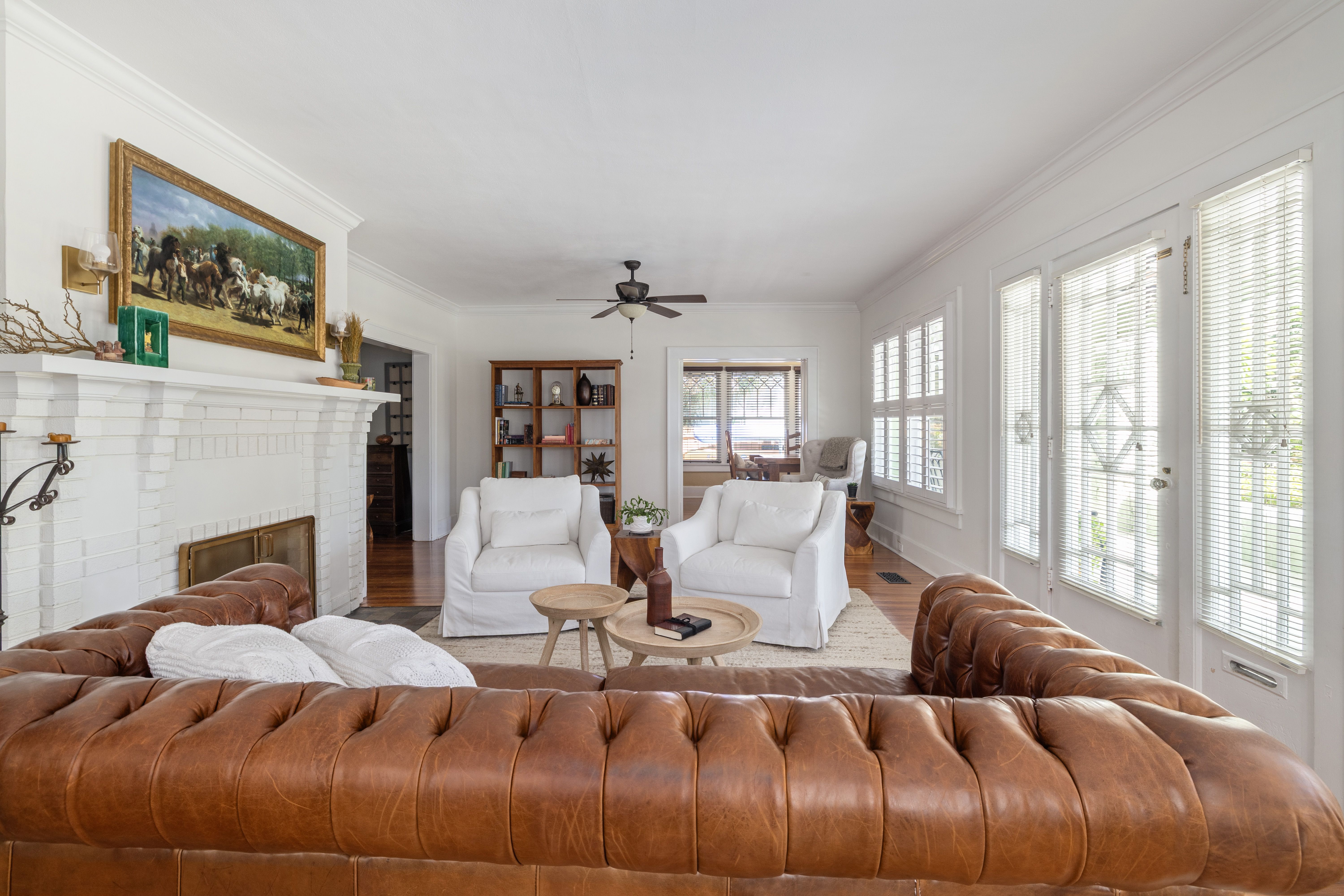 Bright living room with white walls, brown leather couch, two white armchairs, wooden coffee tables, large windows with blinds, white brick fireplace, and horse painting above mantel.