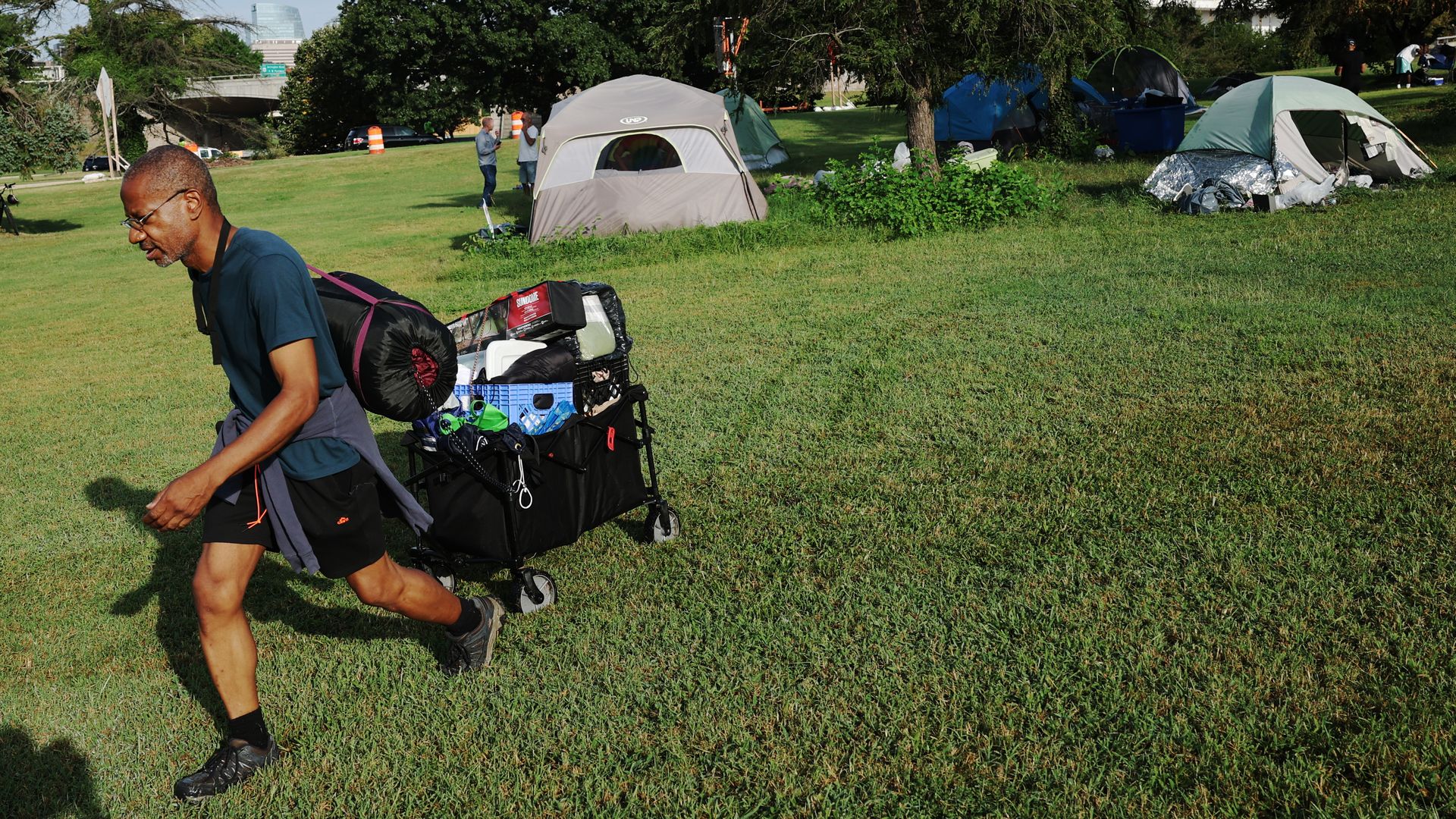 Man in blue shirt and black shorts pulls a cart full of belongings across grassy area with multiple tents and trees in background under a blue sky.
