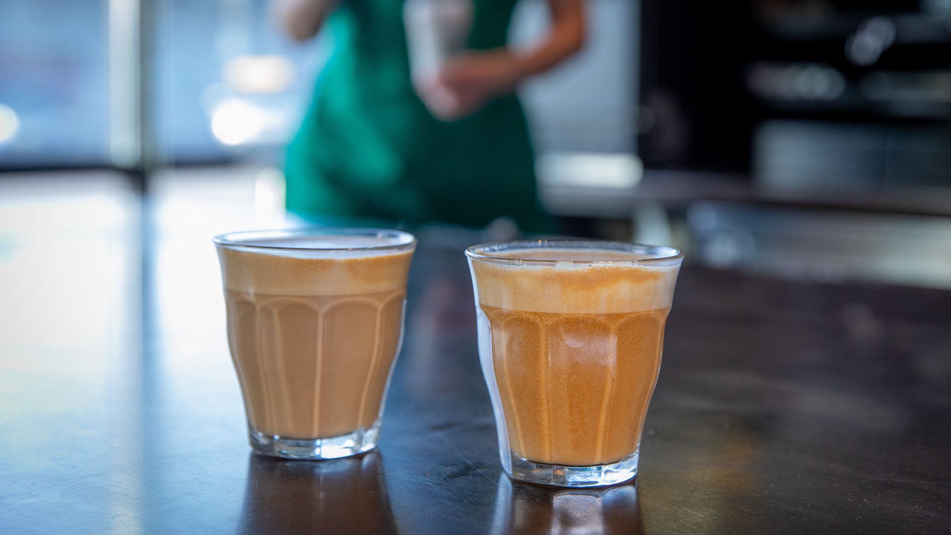 Two glasses with Starbucks Cortado espresso drinks at forefront of photo with a Starbucks barista with green apron in background