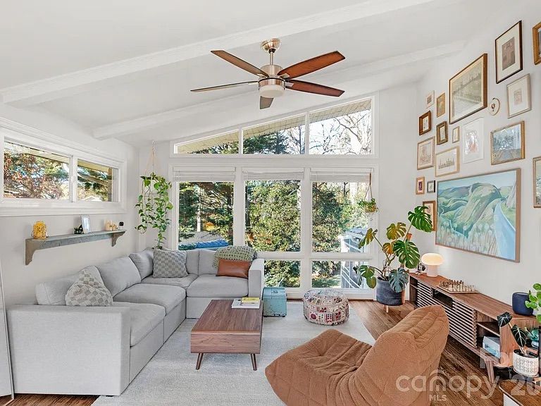 Bright living room with large windows showing greenery, gray sectional sofa, wooden coffee table, brown cushioned chair, wooden media console, plants, and gallery wall of framed artwork.