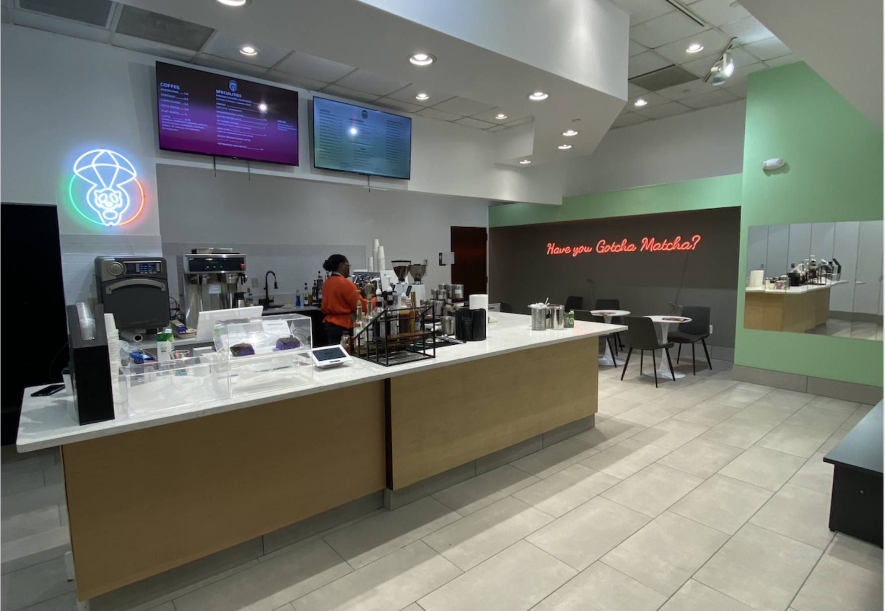 Modern cafe interior with white and wood counter, barista in orange shirt, neon sign reading "Have you Gotcha Matcha?", two digital menu screens, and green accent wall.
