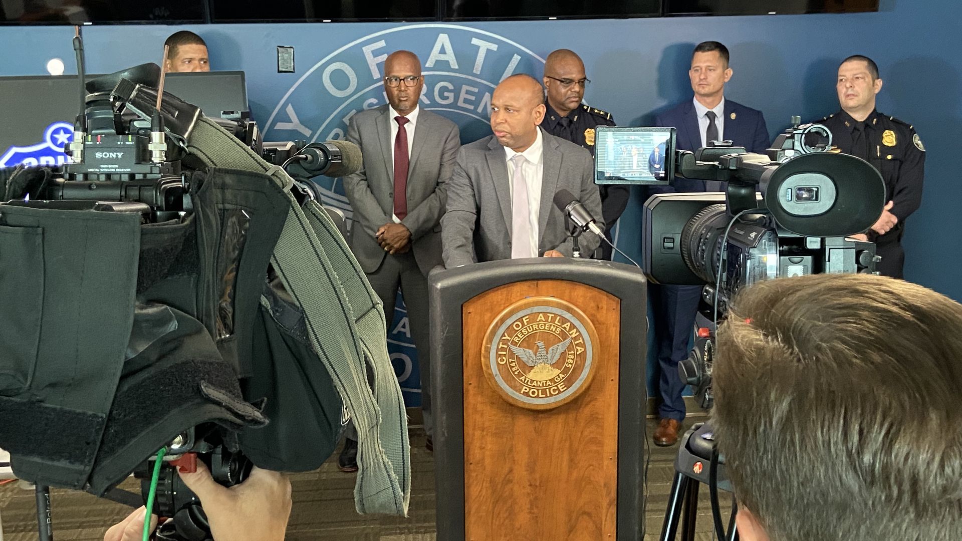 A detective wearing a suit stands at a lectern flanked by police officers
