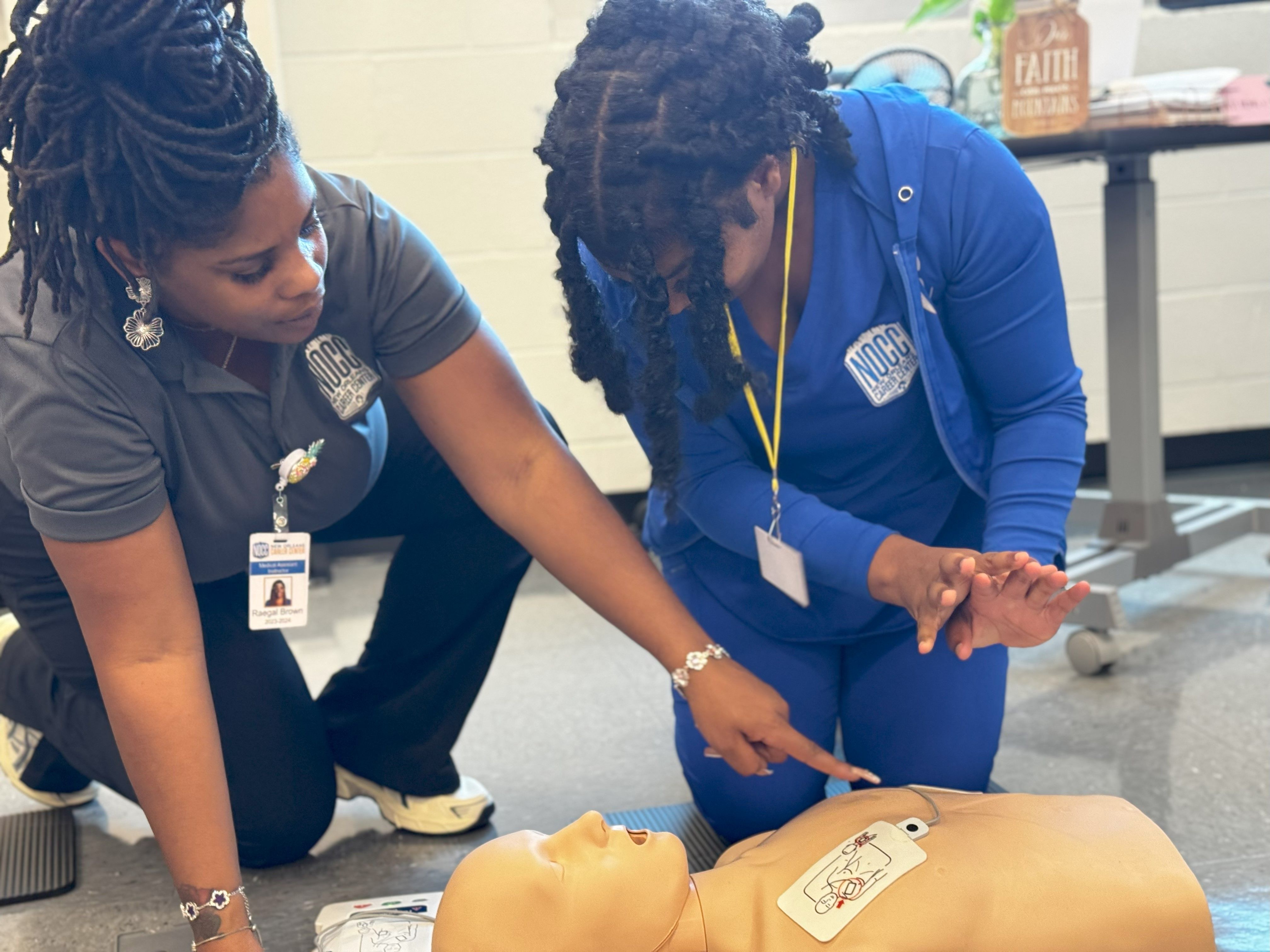An instructor gestures toward a torso dummy on the floor while a student looks on, prepared to perform CPR.