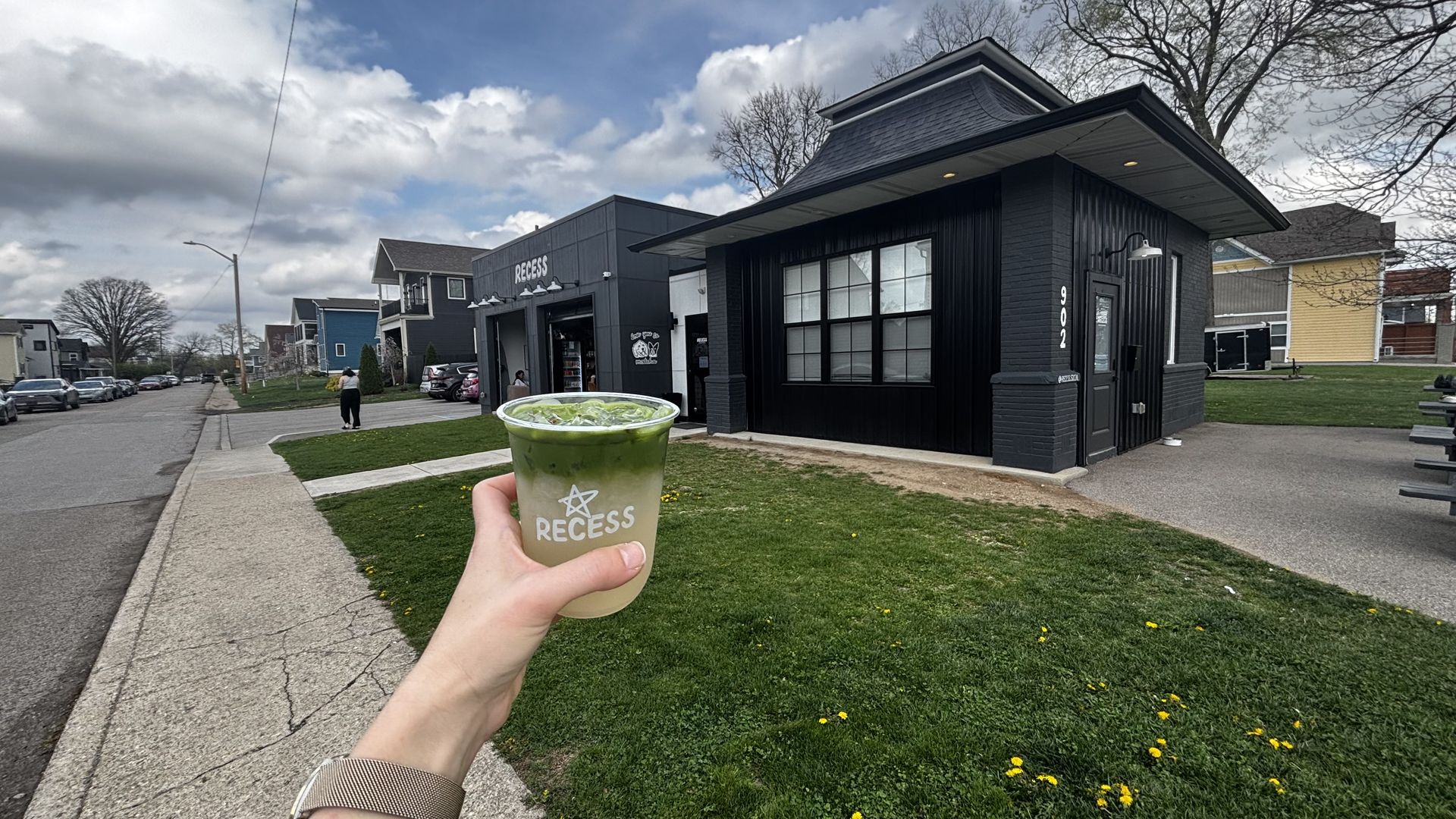 First-person view of a hand holding a clear cup of iced matcha from RECESS in front of a dark storefront on a residential street; cloudy sky, green lawn, parked cars, and a person walking.