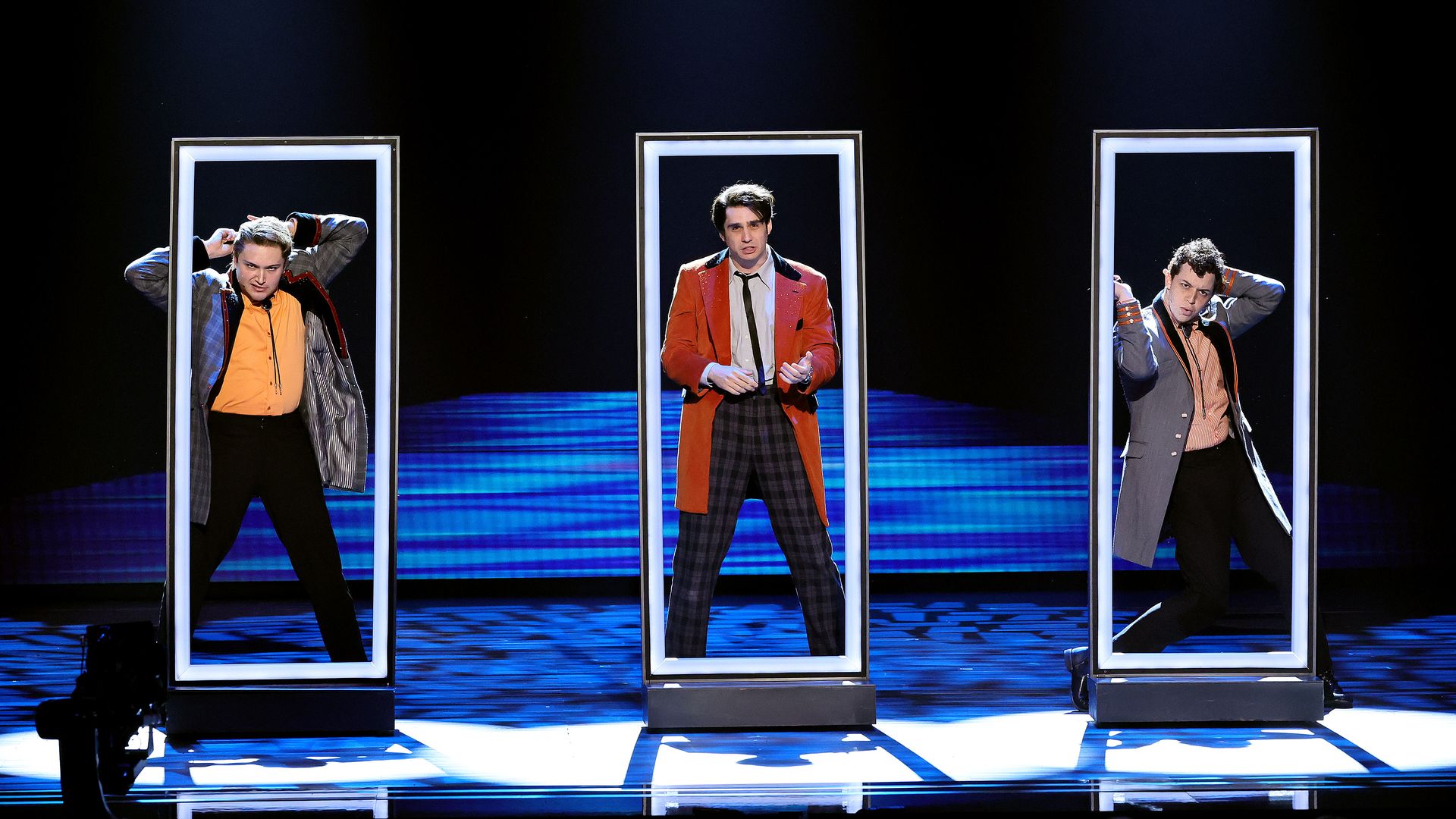 Three men on stage each standing inside a tall rectangular frame under spotlights, dressed in colorful retro-style jackets and pants, with a dark blue background.