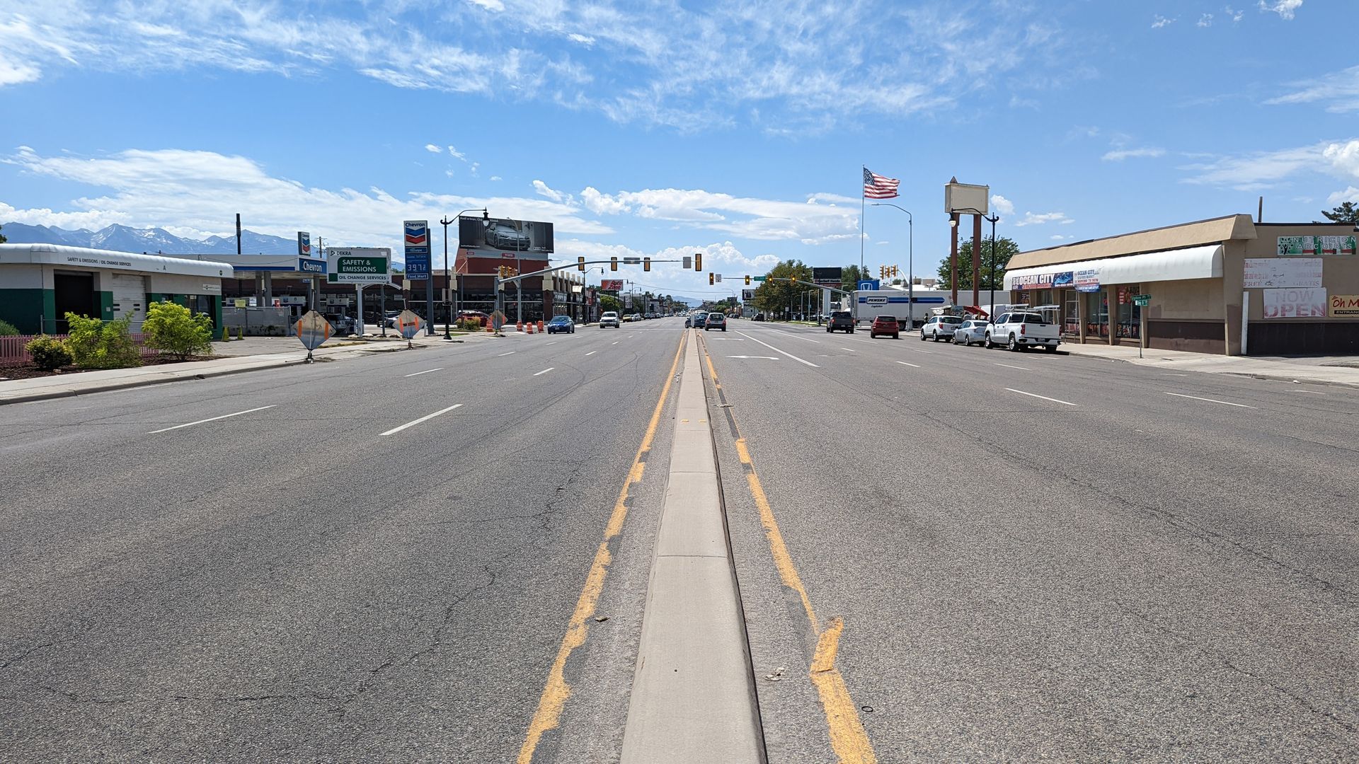 A six-lane road with a narrow paved median and a few buildings and cars.