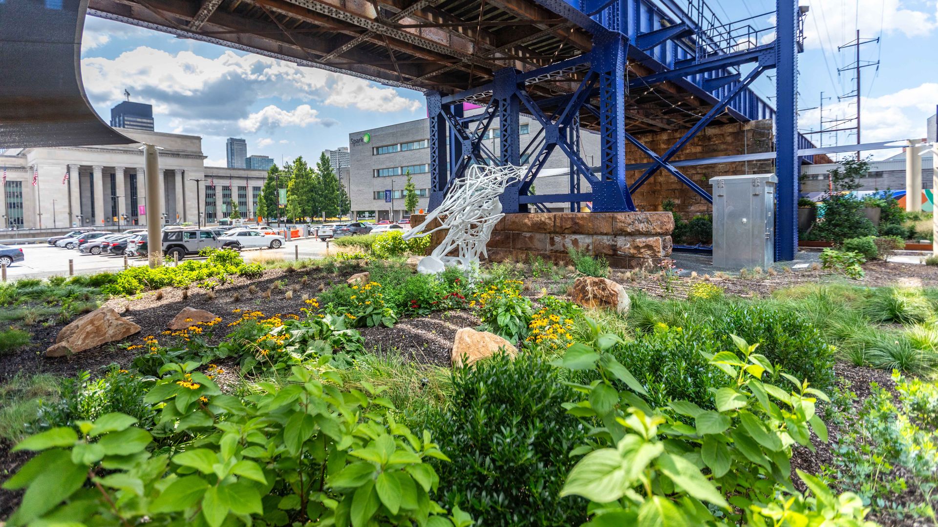 Highland Park in University City beneath a blue steel rail overpass, which includes leafy green plants and yellow flowers, white abstract sculpture, parked cars, and city buildings under a partly cloudy sky.