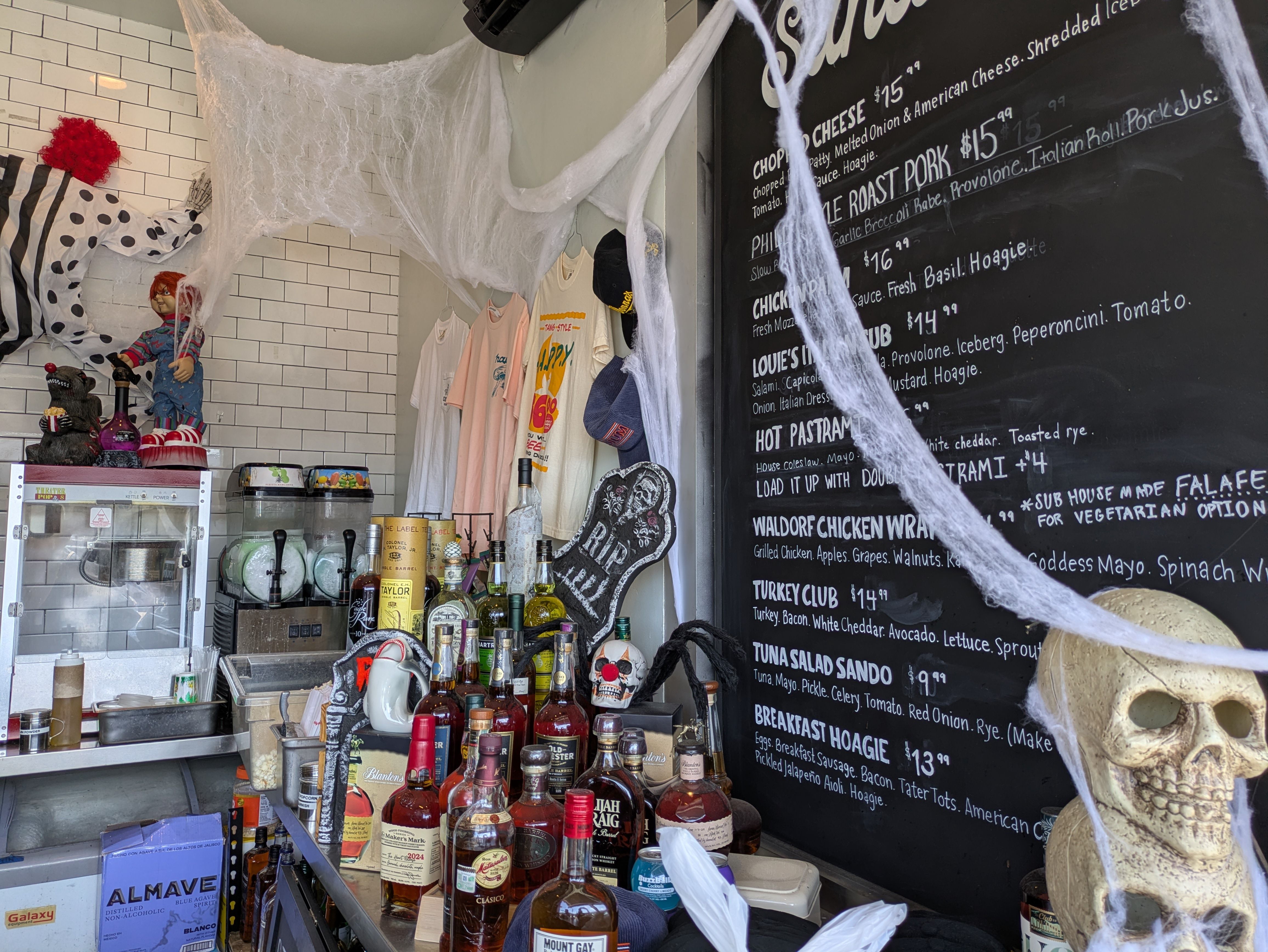 Bar corner with Halloween decorations including fake cobwebs and a skull, liquor bottles, a popcorn machine, T-shirts hanging on wall, and a blackboard menu listing sandwiches and prices.