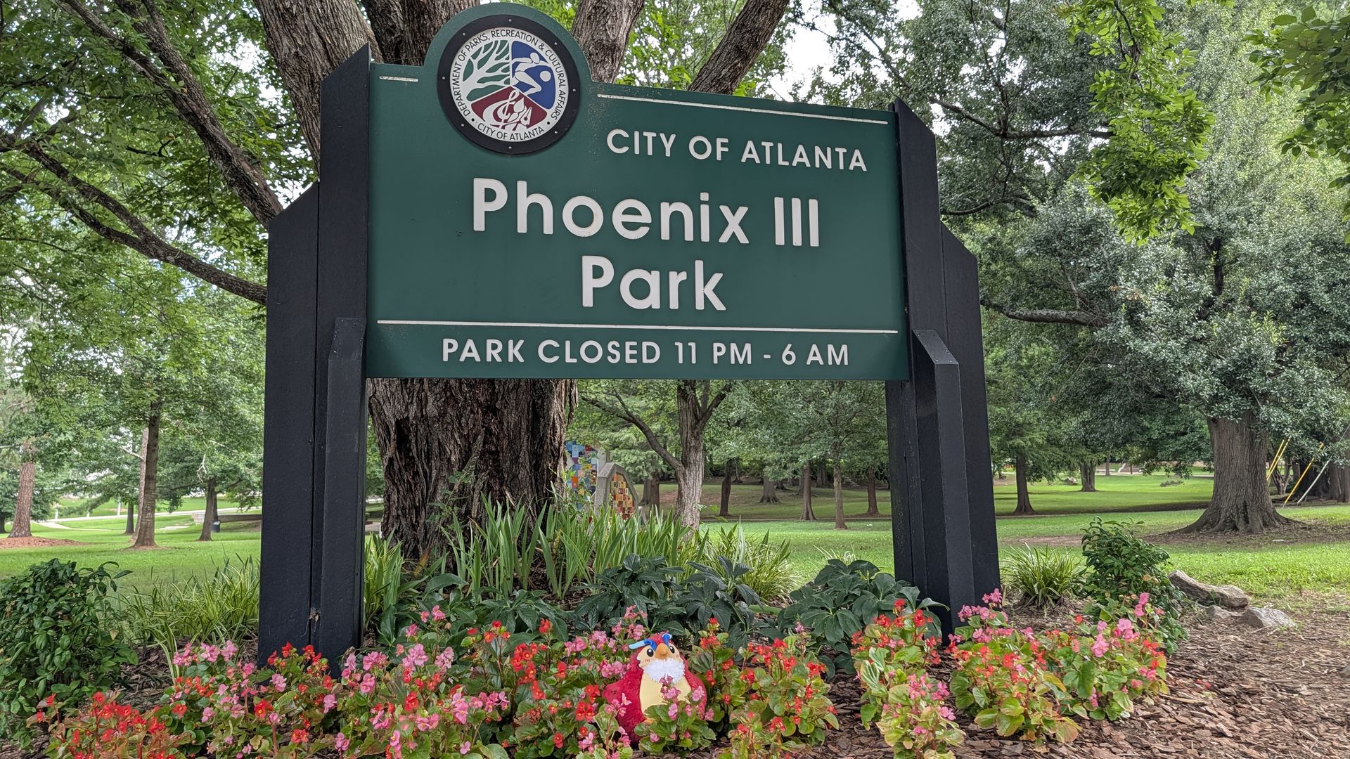 Green park sign reading "City of Atlanta Phoenix III Park" with a circular seal at the top, surrounded by trees. In front, pink flowers and greenery; hours read "Park closed 11 PM - 6 AM".