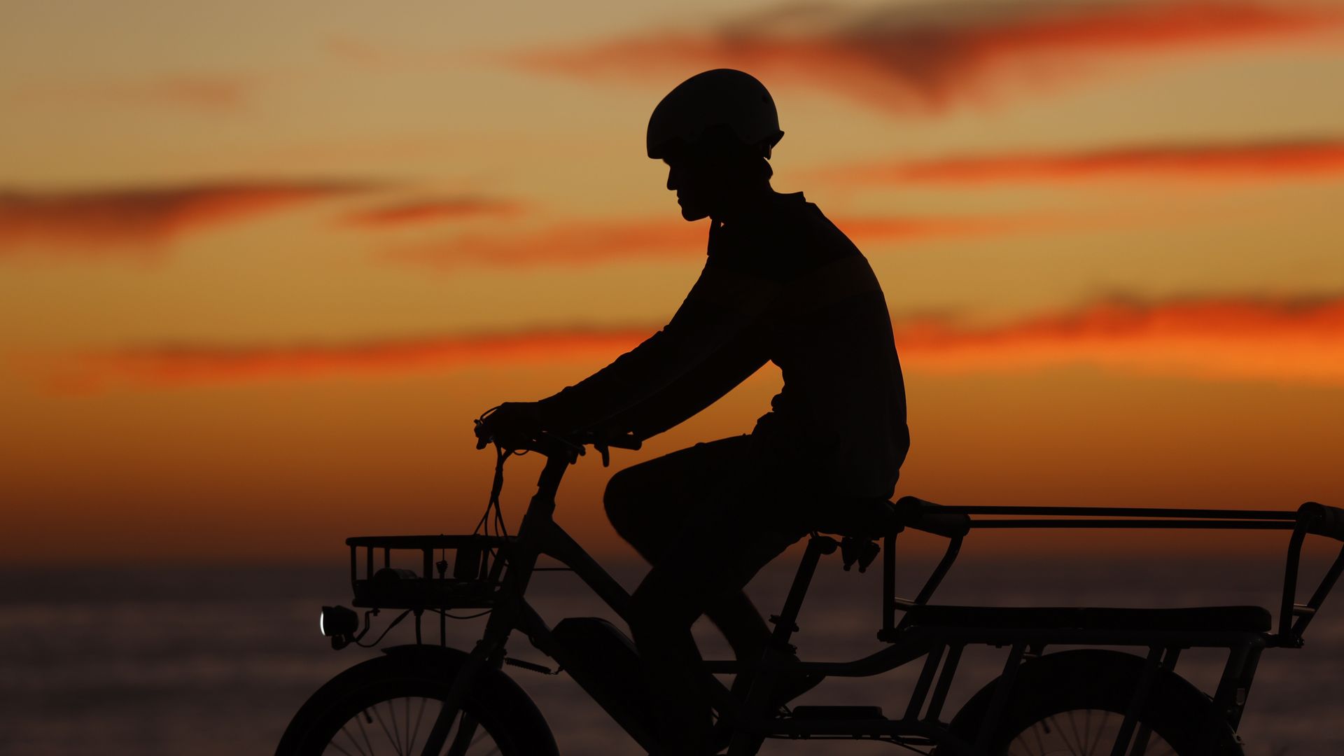 Silhouette of a boy wearing a helmet riding a cargo-style e-bike along a waterfront at sunset, with vivid orange and red clouds streaking across the sky behind them.