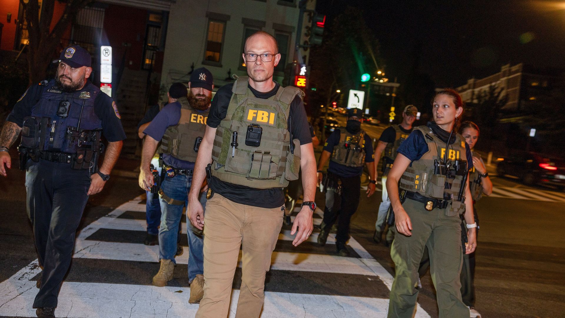 A group of FBI and law enforcement officers wearing tactical vests walk on a city street at night with buildings and traffic lights visible in the background.