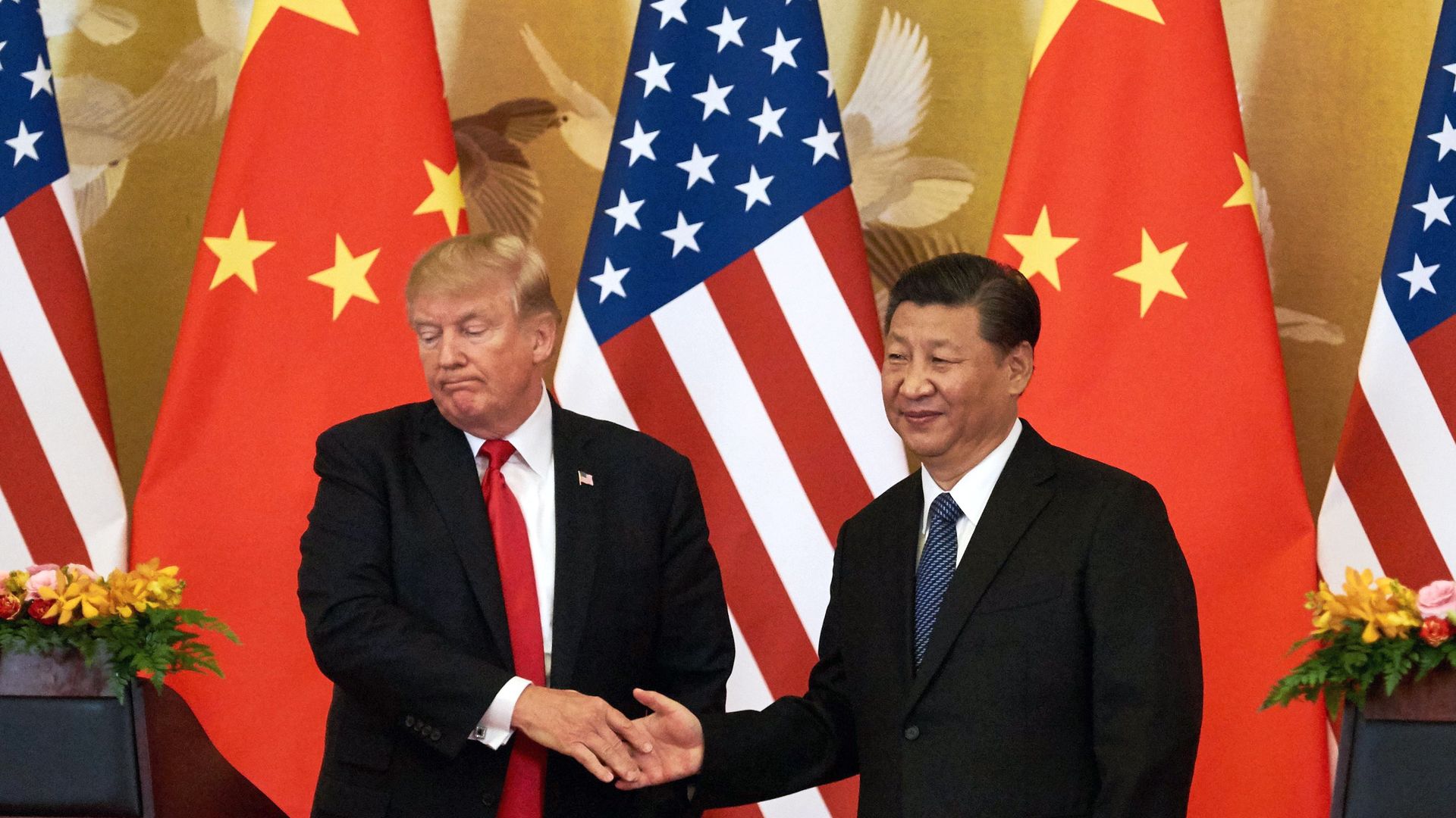 Donald Trump grimaces while shaking hands with Xi Jinping before American and Chinese flags.