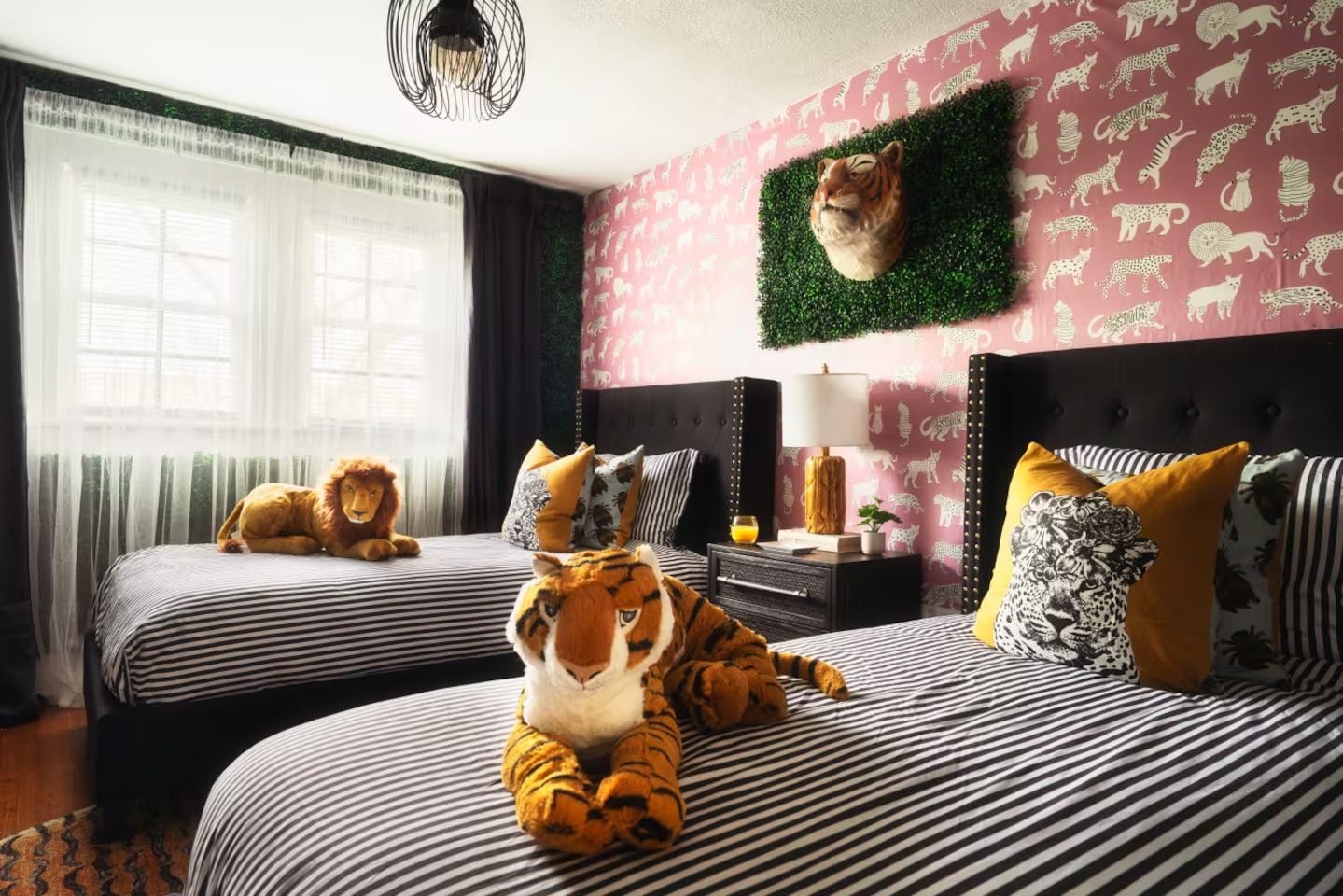 A bedroom with two twin beds with black and white striped bedding, animal-themed pillows, stuffed tiger and lion toys, a tiger head mounted on a green plant wall panel, and pink wallpaper with white animal silhouettes.