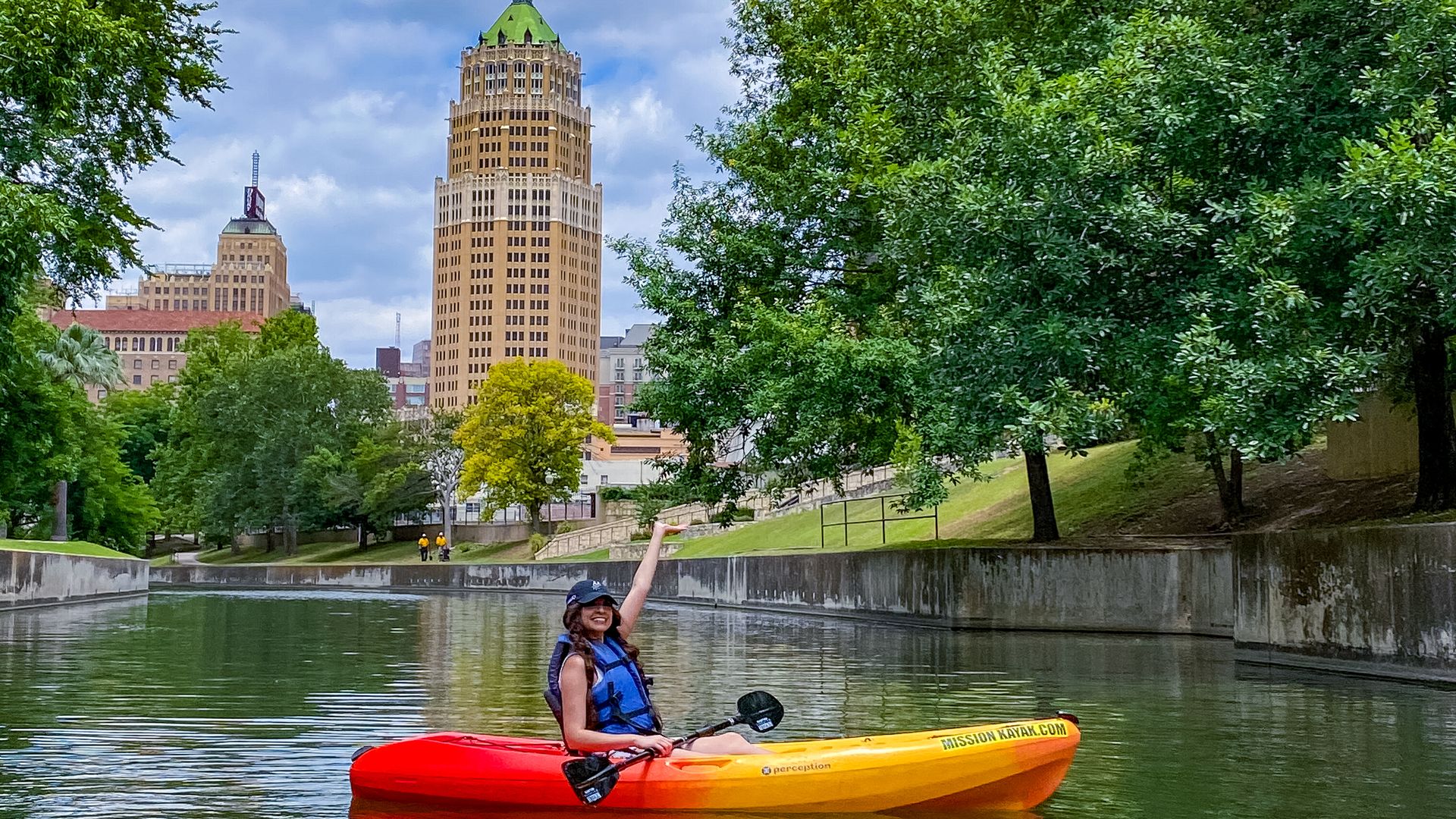 A woman sits in a brightly colored kayak in the middle of the San Antonio River with a slender skyscraper in the background.