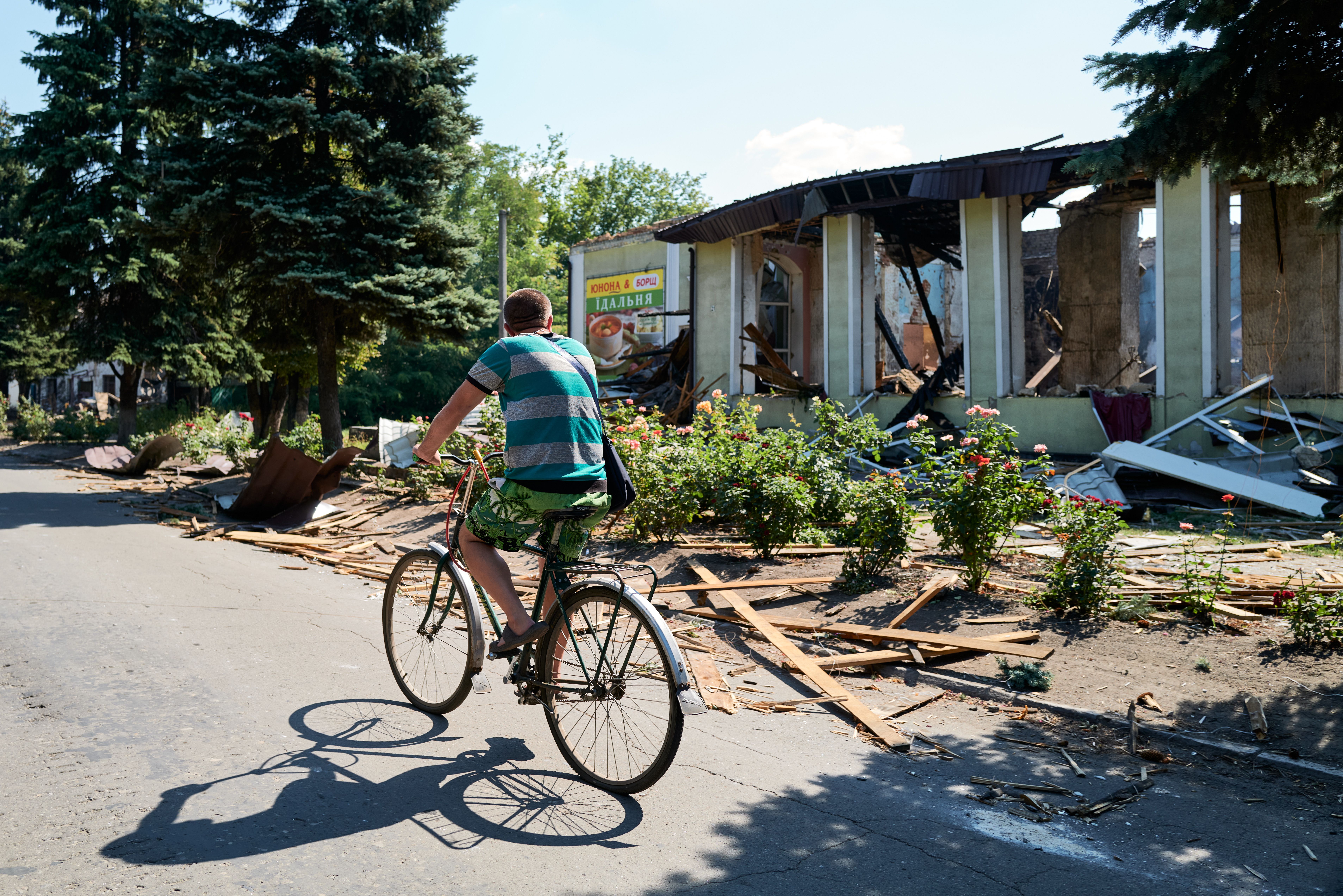 Man in green striped shirt and shorts rides bicycle on road past damaged building with debris and rose bushes in sunlight under blue sky.