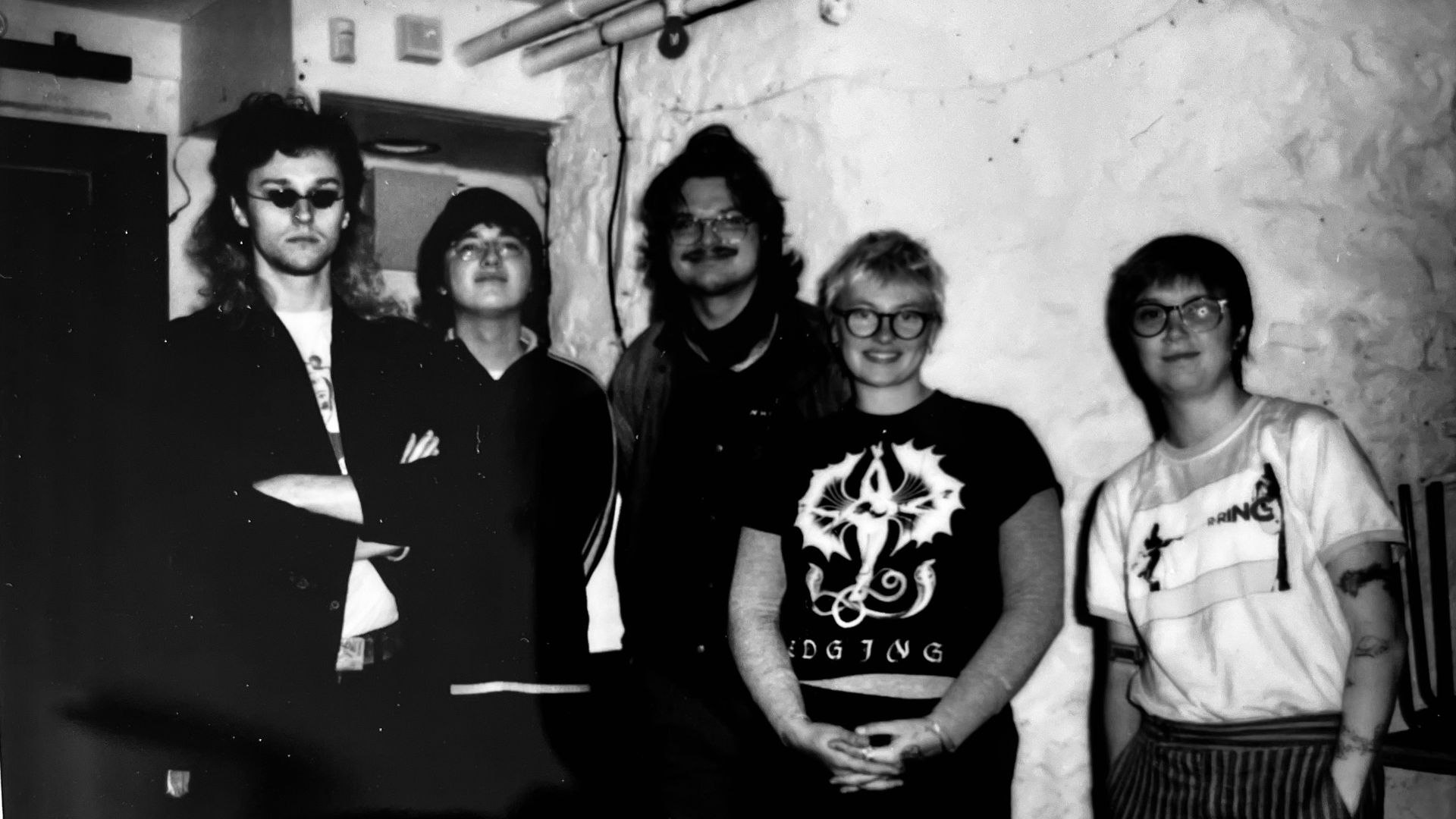 Black-and-white photo of five people posing in a basement with exposed pipes. Left to right: man with sunglasses and long hair; others wearing glasses; a woman in a graphic tee; a rightmost person in striped pants.