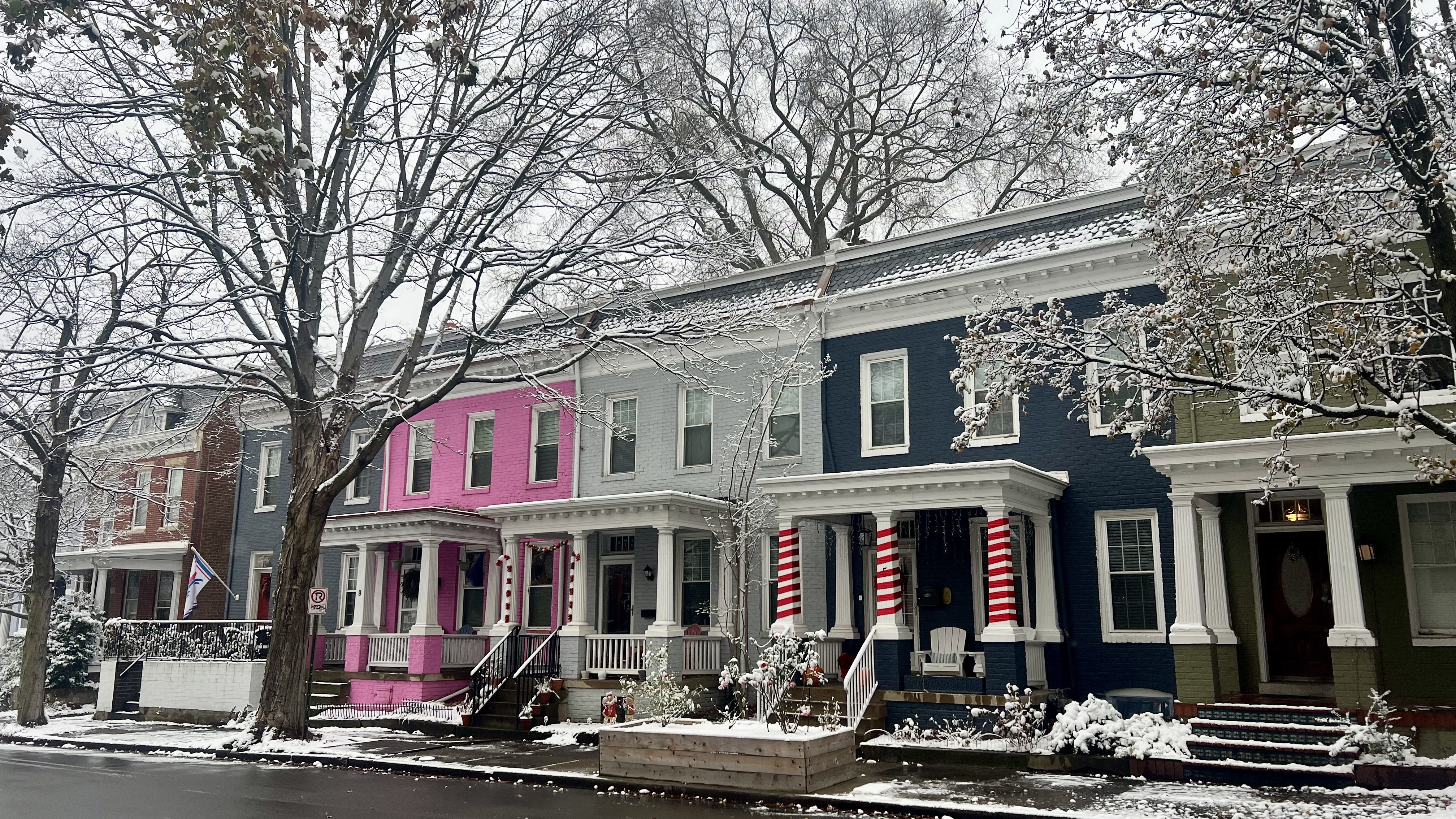 Snow-covered street with colorful row houses in blue, pink, gray, and green. Trees with snow on branches line the sidewalk and holiday candy cane decorations adorn a porch.