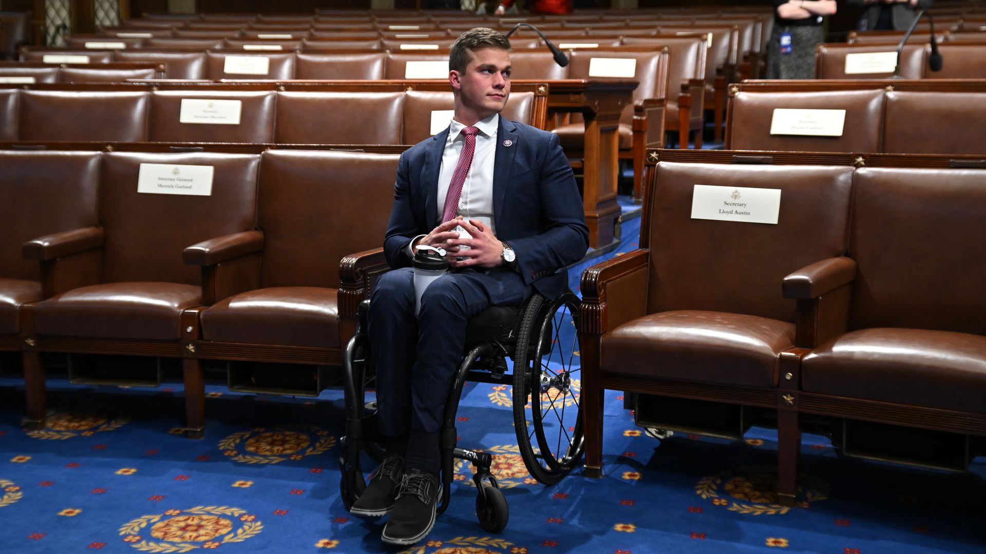 A man in a wheel chair on the floor of the House of Representatives wearing a blue suit and pink tie, looking off to the side 