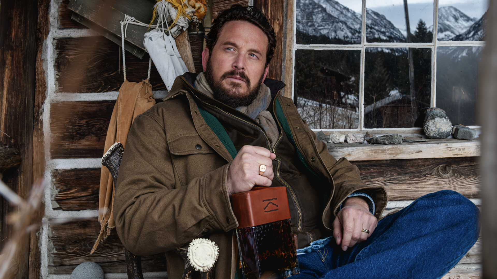Actor Cole Hauser with the Lazy K Bar bourbon, sitting on a chair in a coat and jeans.