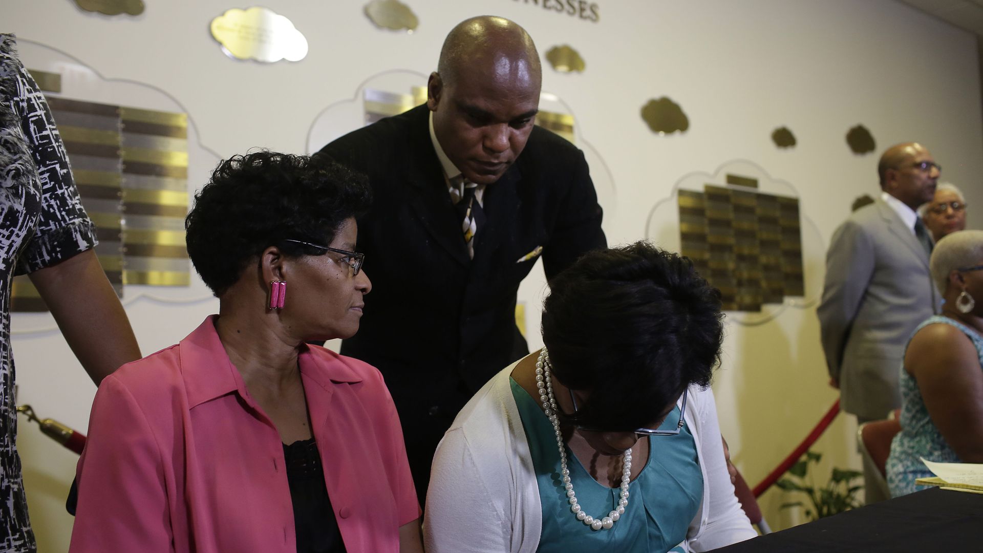 In this image, a man in a suit leans over two seated woman at a table with a black tablecloth.
