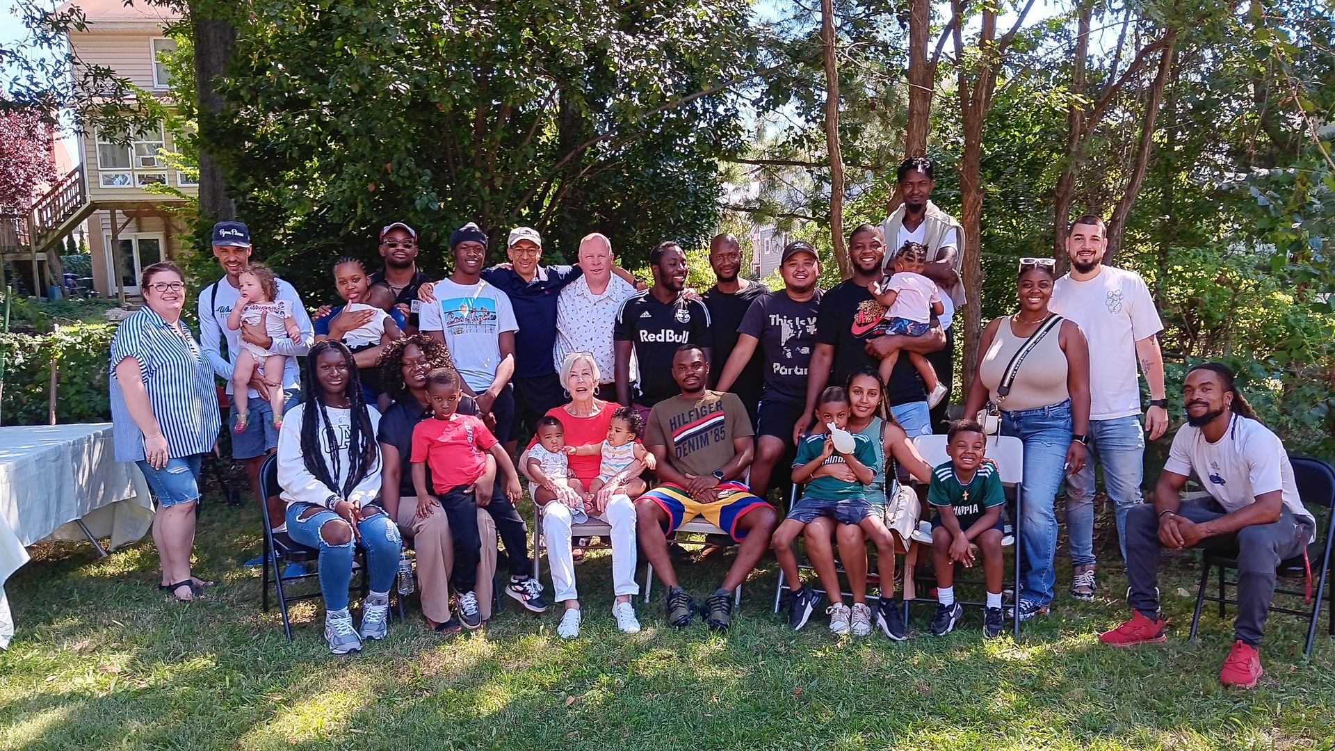 Large group of adults and children posing for a sunny outdoor group photo in a backyard with trees and a house in the background; everyone smiles in casual clothing.