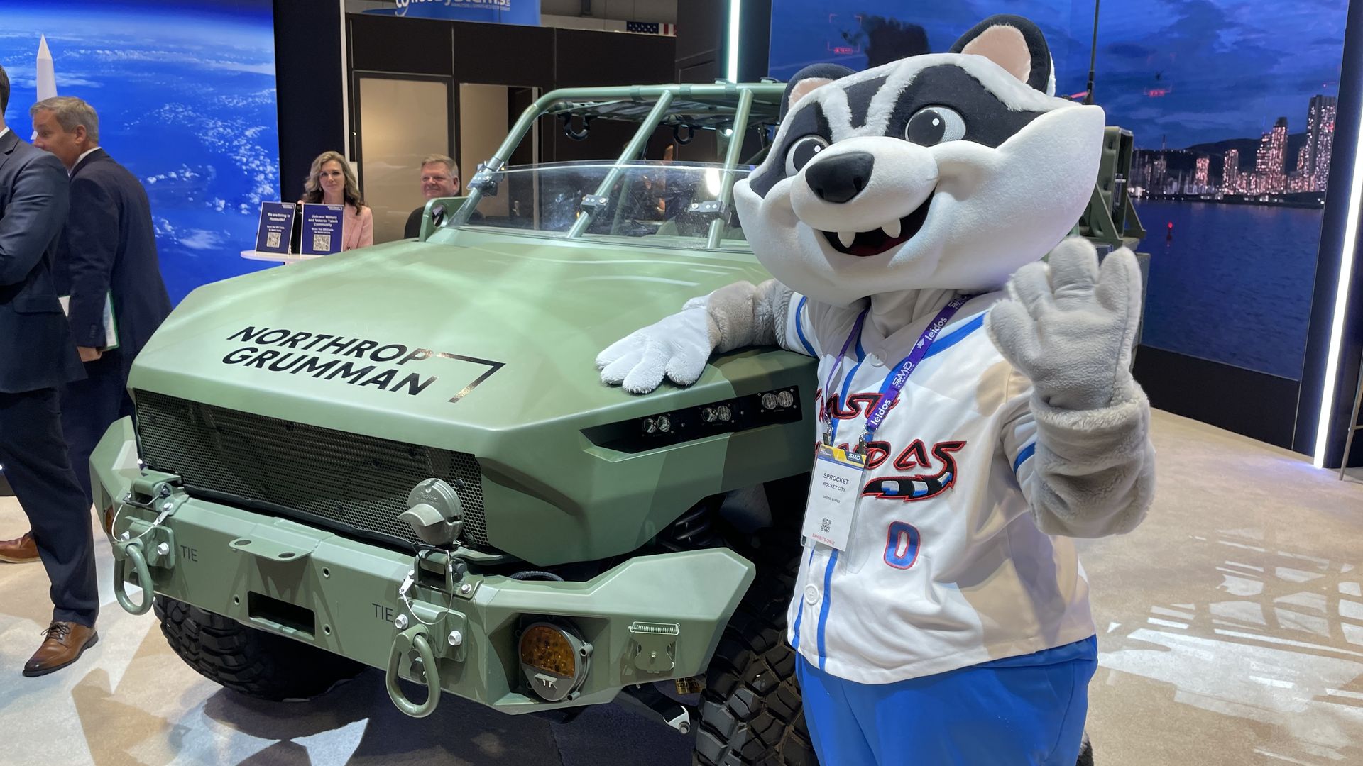 At an expo, a light-green military-style vehicle labeled Northrop Grumman sits beside a large raccoon mascot in a white suit waving. People in business attire stand against a cityscape backdrop.