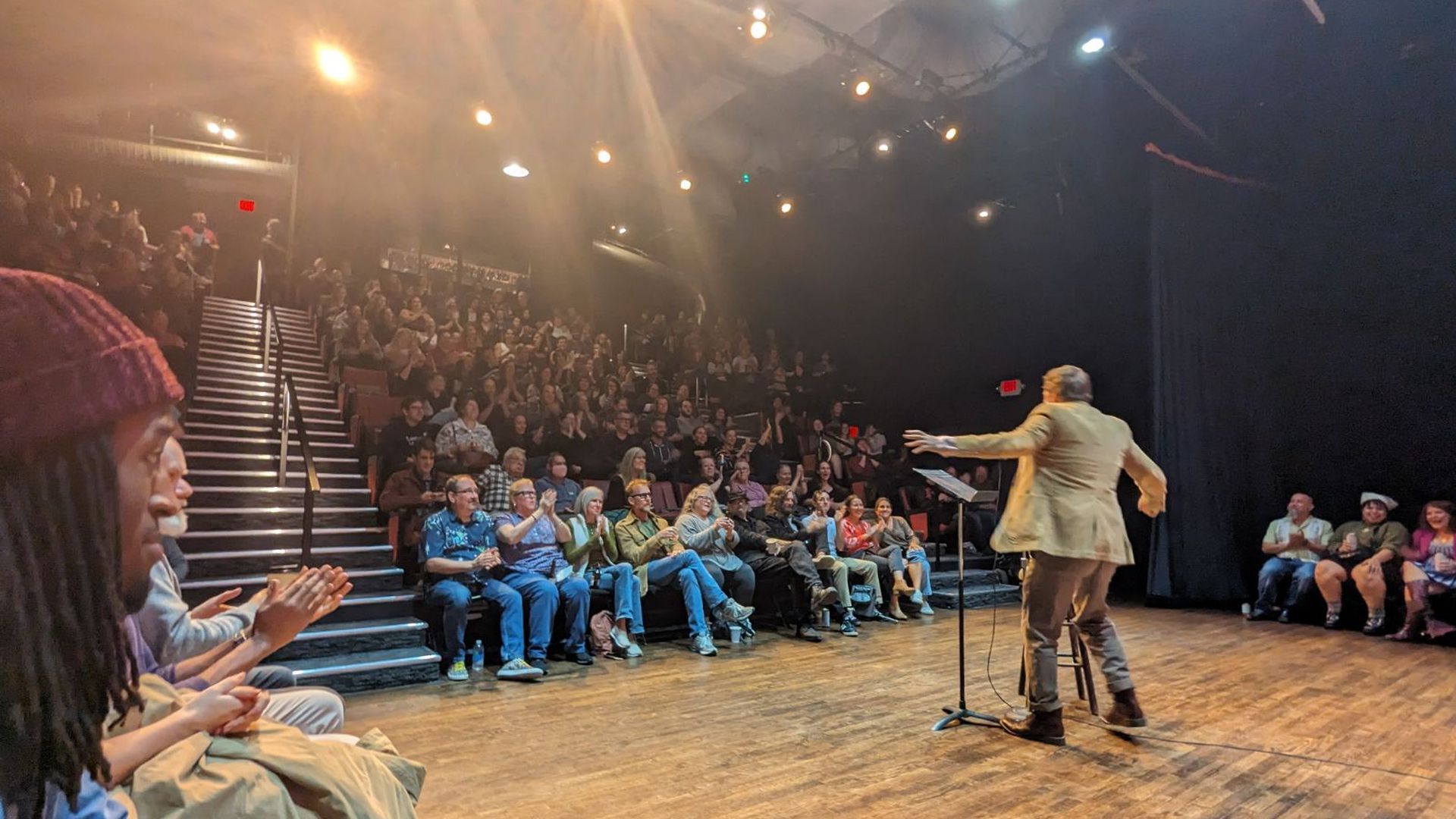 A man in a coat jacket stretches out his arms while telling a story to a bright packed theater with stadium-style seating