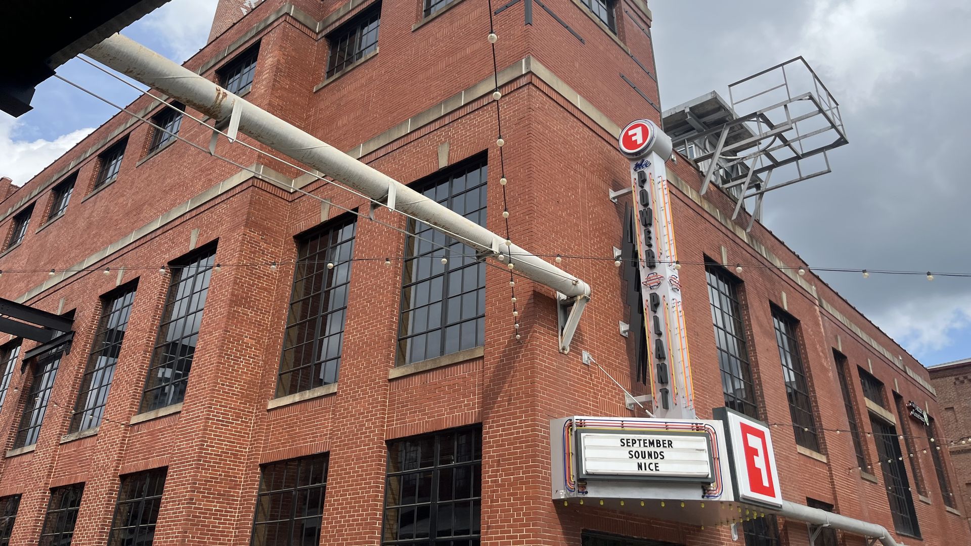 Red brick industrial building with large grid windows, gray stone trim, and string lights. A tall corner tower rises under a cloudy sky; a vertical sign and marquee read "September Sounds Nice".