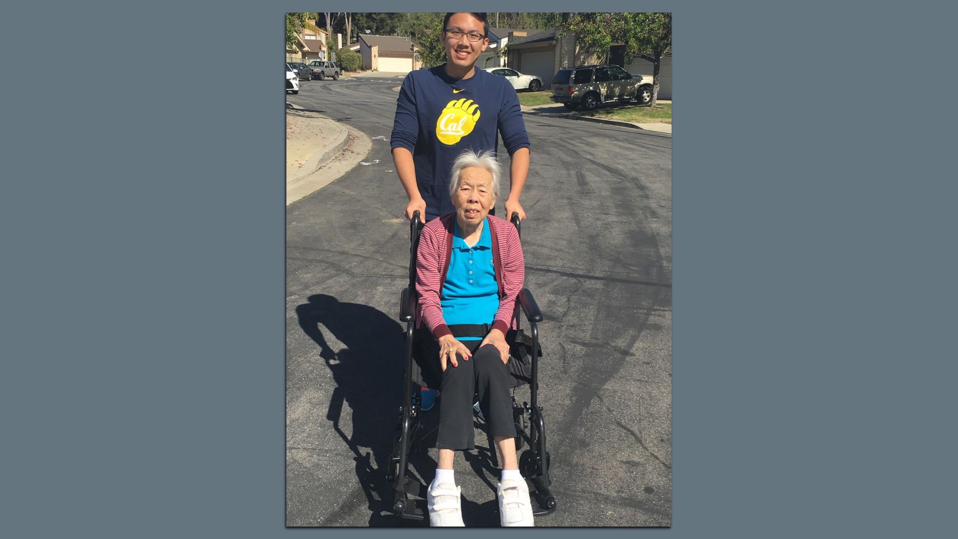Denny Lai wheeling his grandmother, Bao Ling, through a residential street with parked cars and houses.
