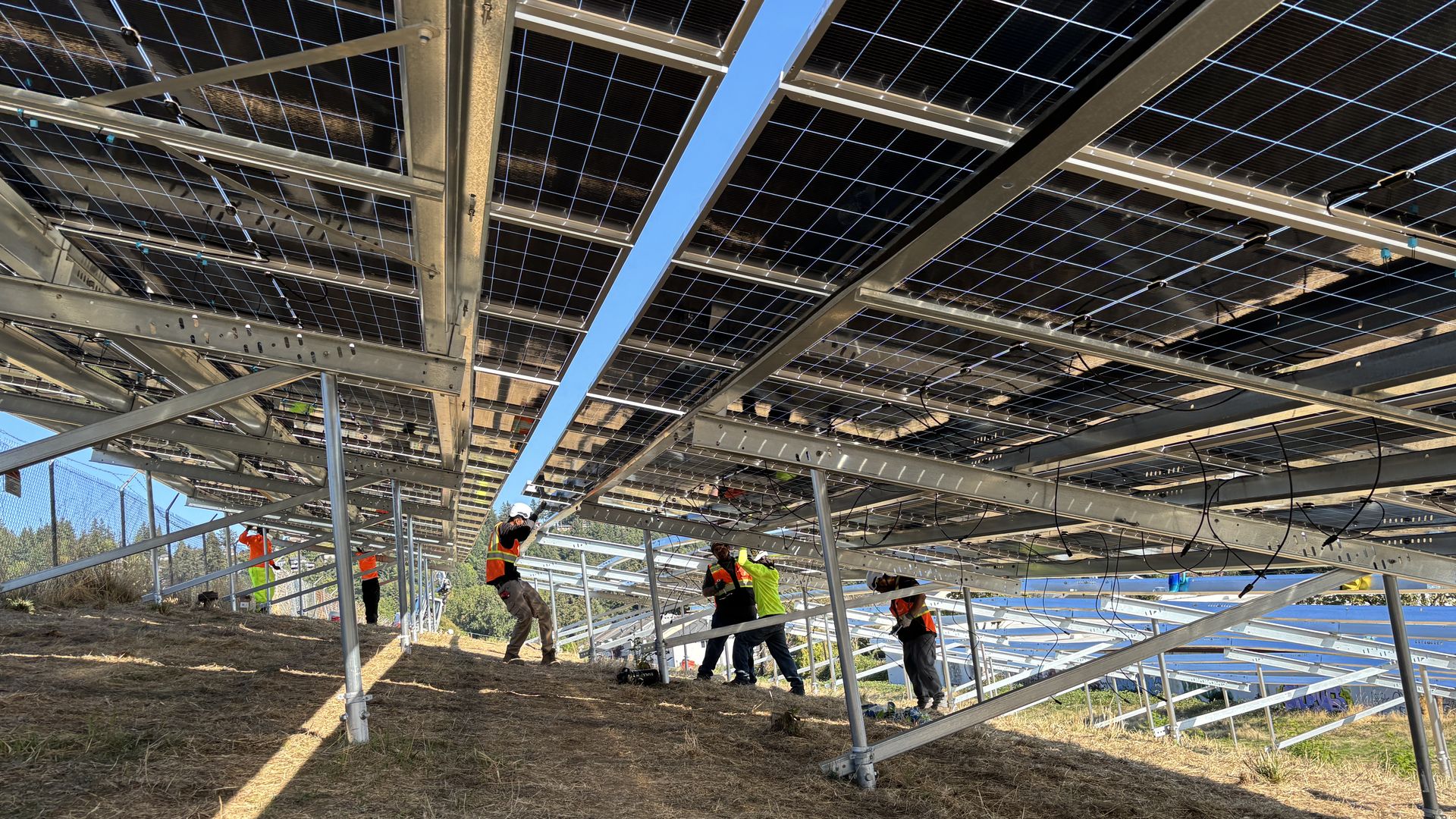 Construction workers in orange vests install and inspect metal frames supporting numerous solar panels mounted overhead, with blue sky visible through the grid and dry grassy ground below.